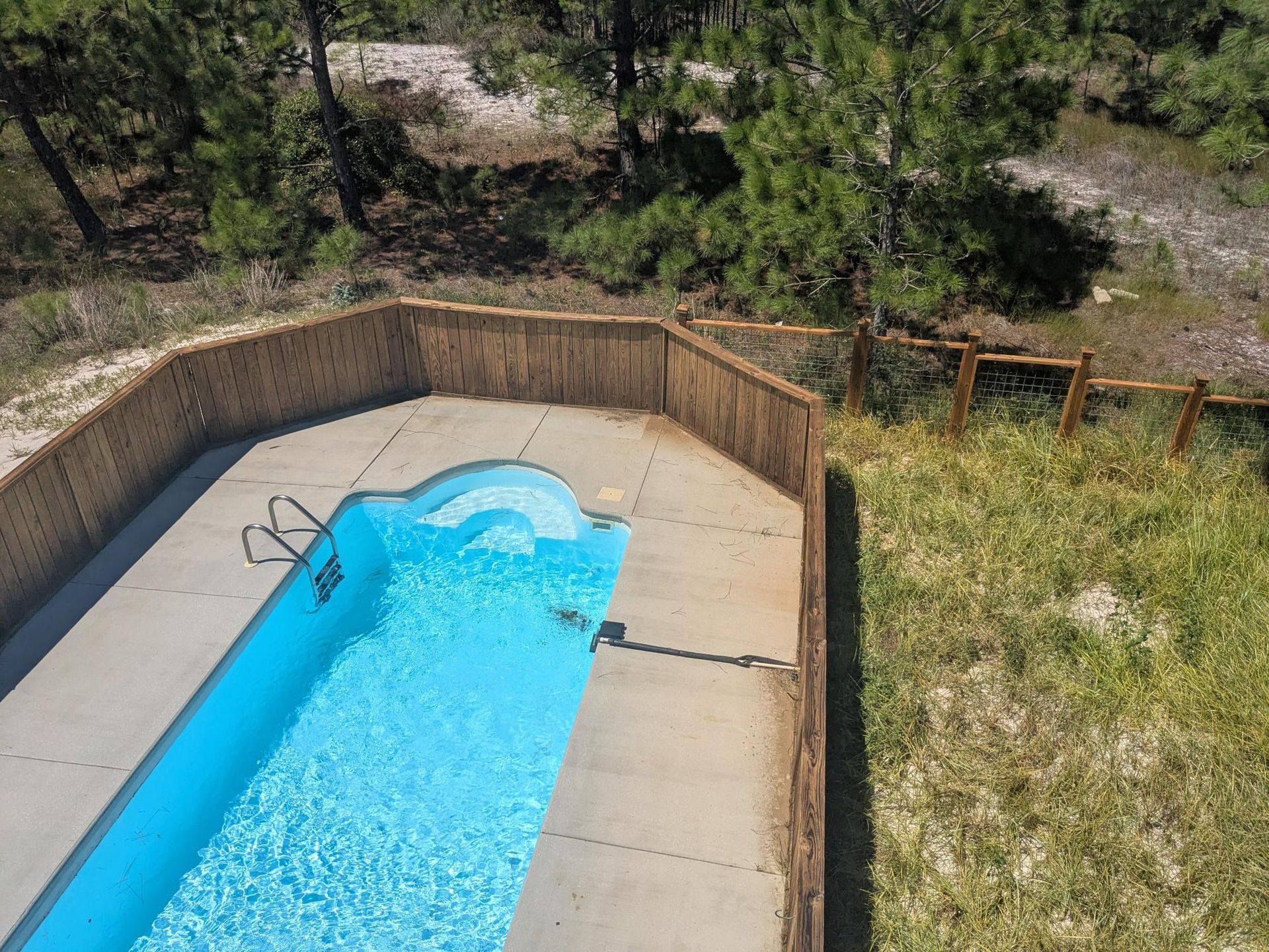 Pool surrounded by a wooden fence, set in a natural, wooded area. Blue water in the pool.