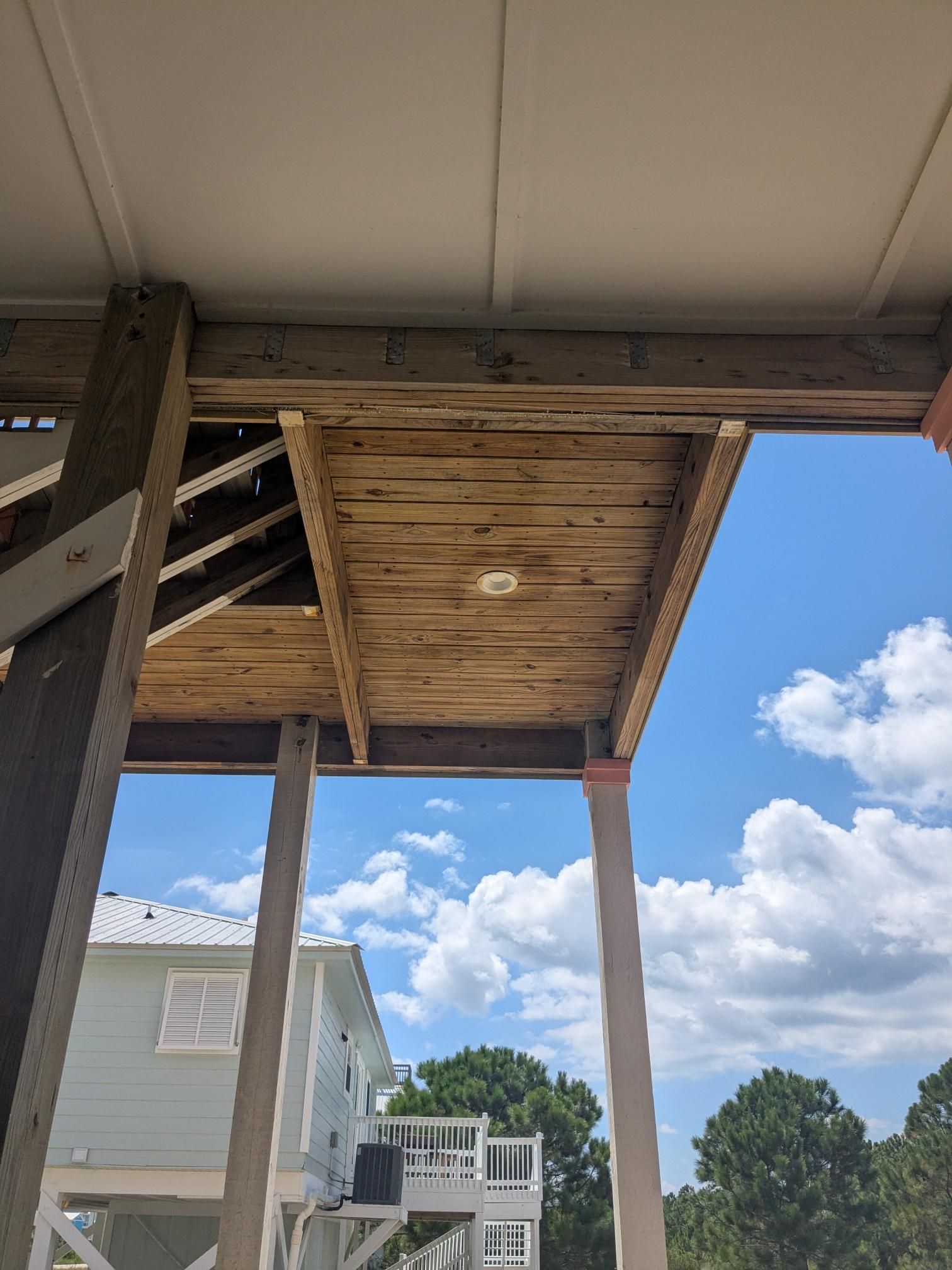 View from under a raised wooden deck with weathered planks. The sky and two houses are visible in the background.