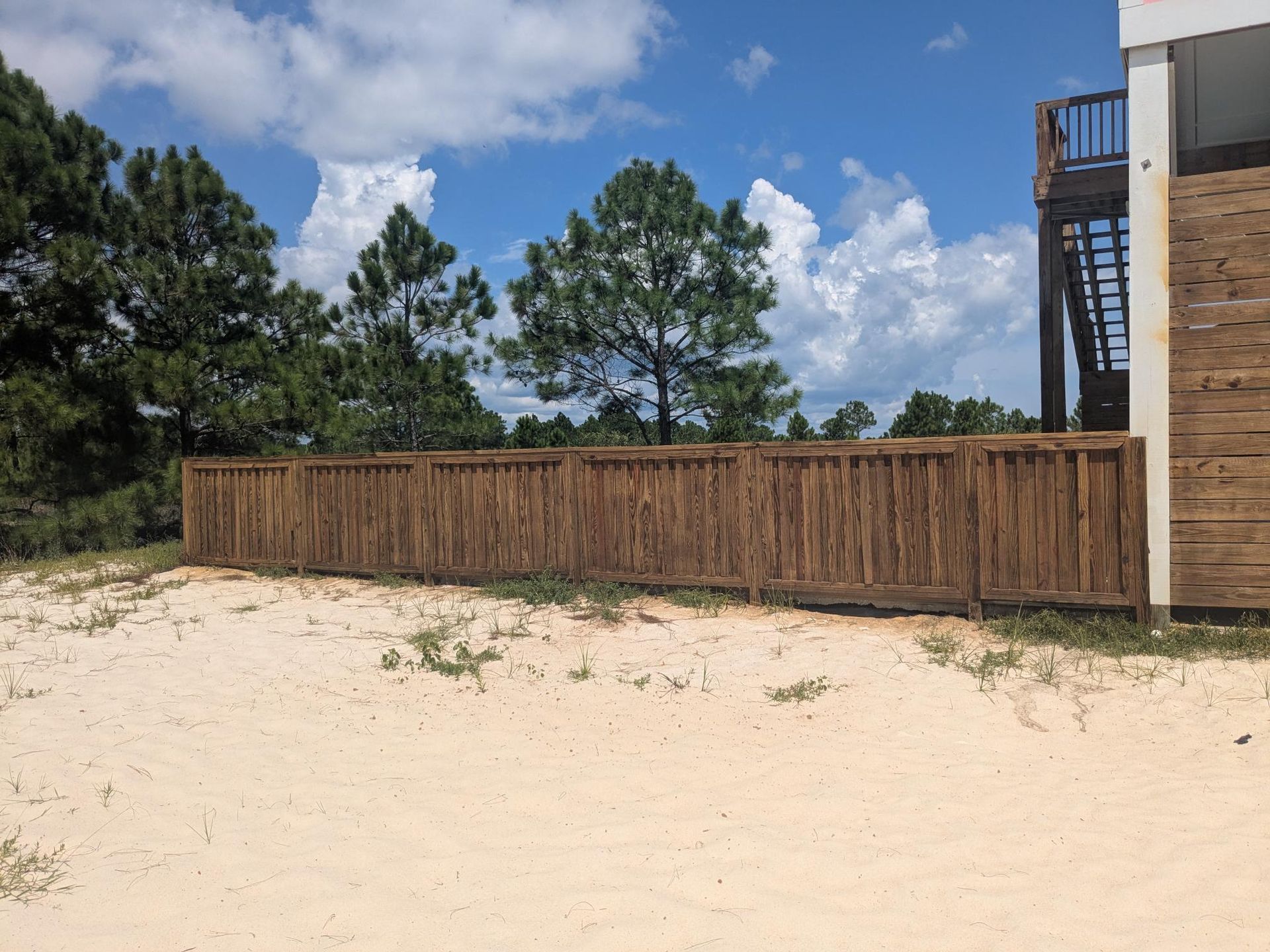 Wooden fence on sandy ground with trees and a building against a blue sky.