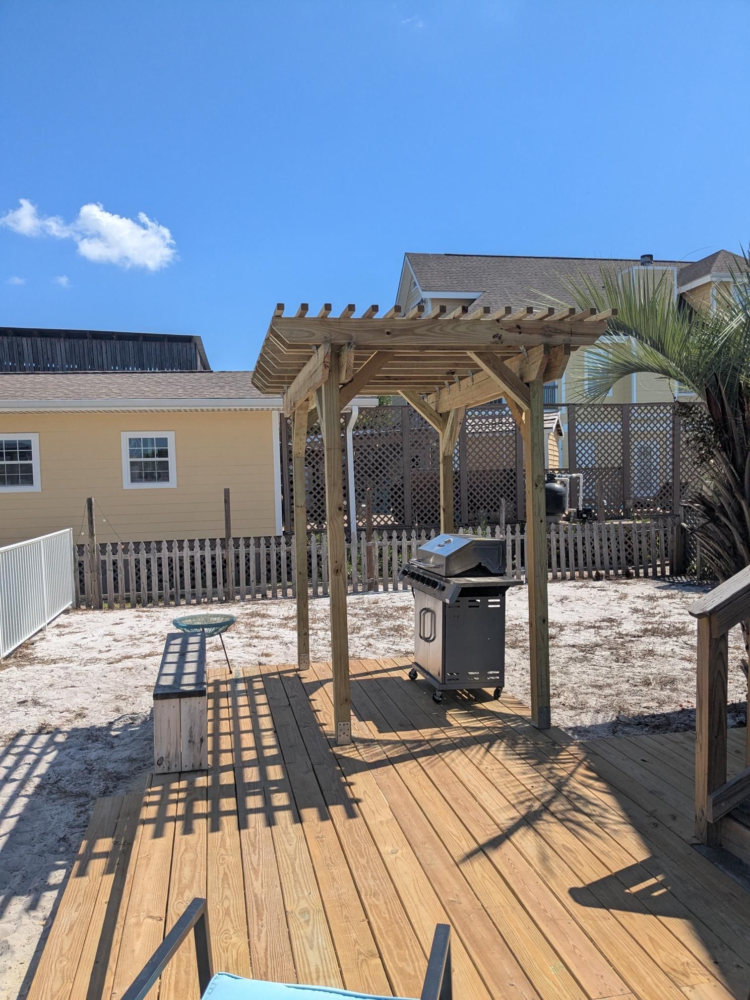 Wooden pergola with grill on a wooden deck in a backyard with a yellow house and fence.