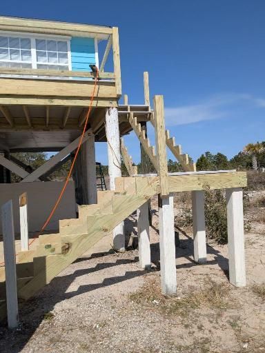 Wooden stairs and deck leading to a house, supported by concrete pillars. Sunny day.