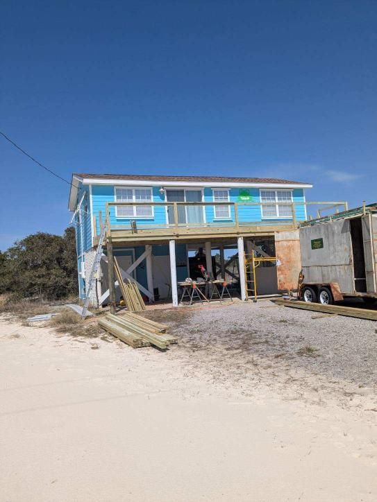 Beach house under construction; light blue siding, wooden deck, poles and trailer on sand, clear sky.