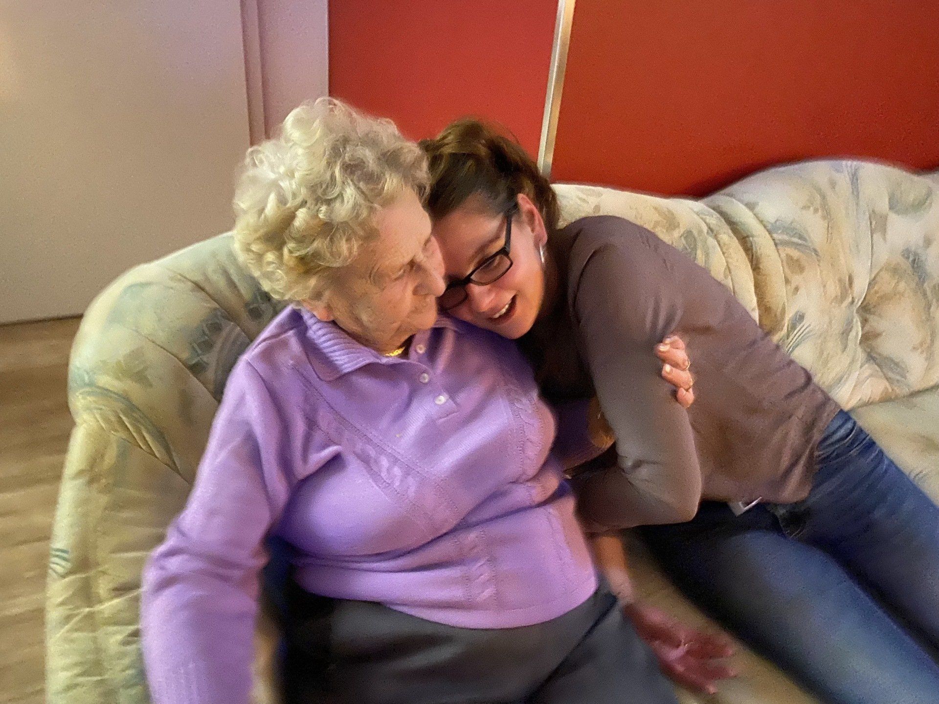 Frau umarmt ältere Person auf einem gemusterten Sofa; beide lächeln.