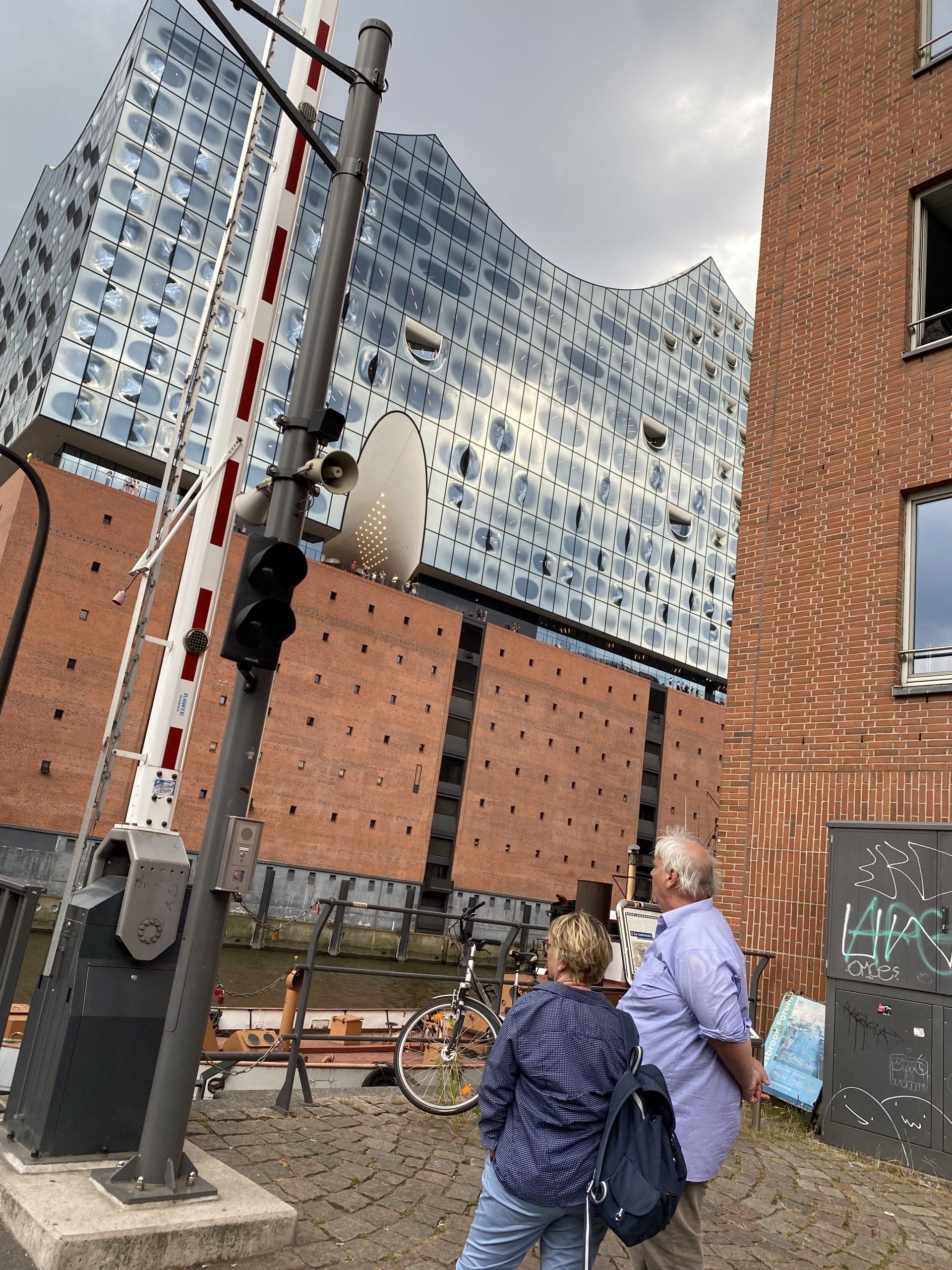 Paar geht am Gebäude der Elbphilharmonie in Hamburg vorbei. Vorne ist eine Schranke.