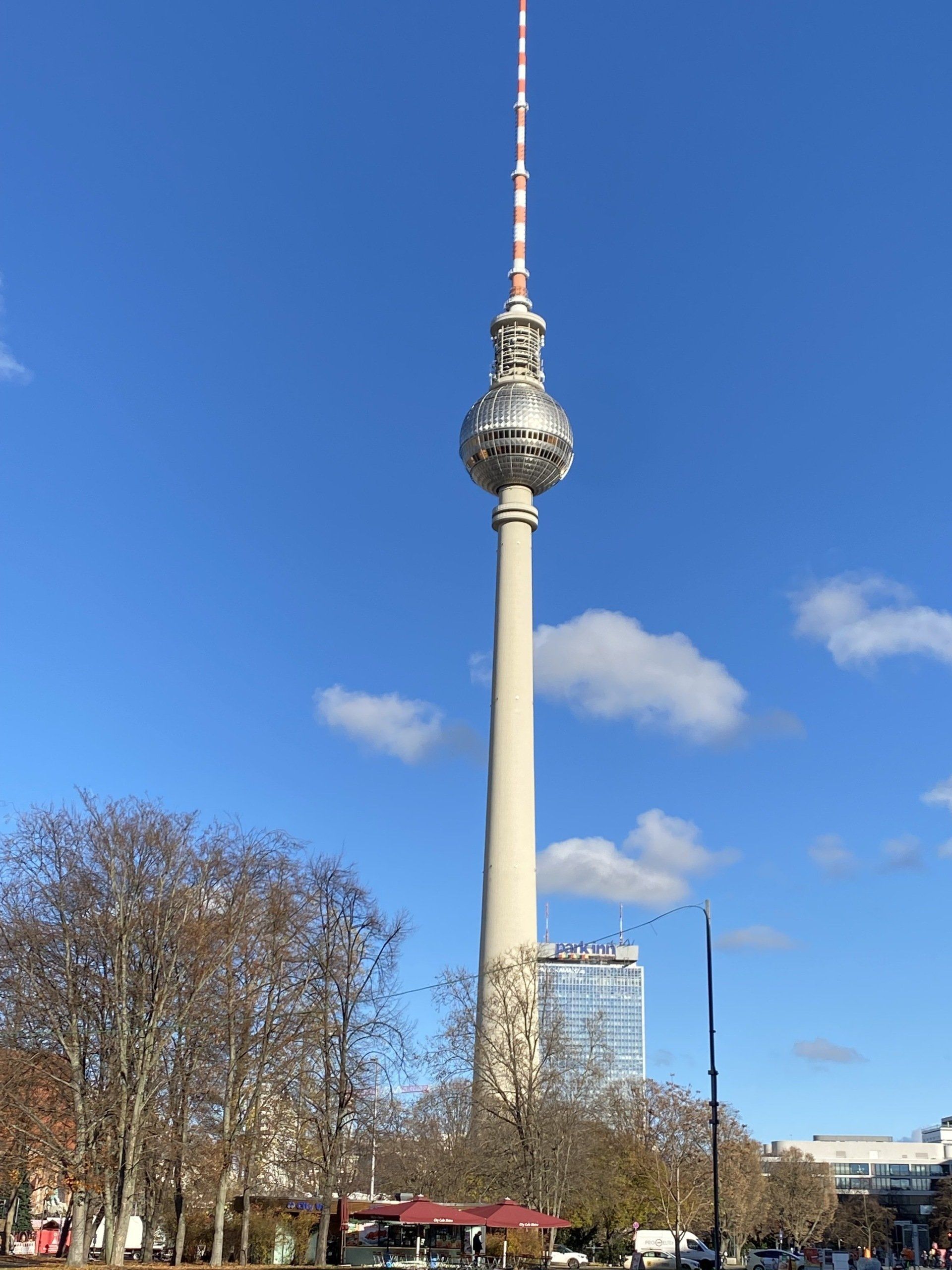 Berliner Fernsehturm vor blauem Himmel, mit einigen Bäumen und einem Gebäude im Vordergrund.