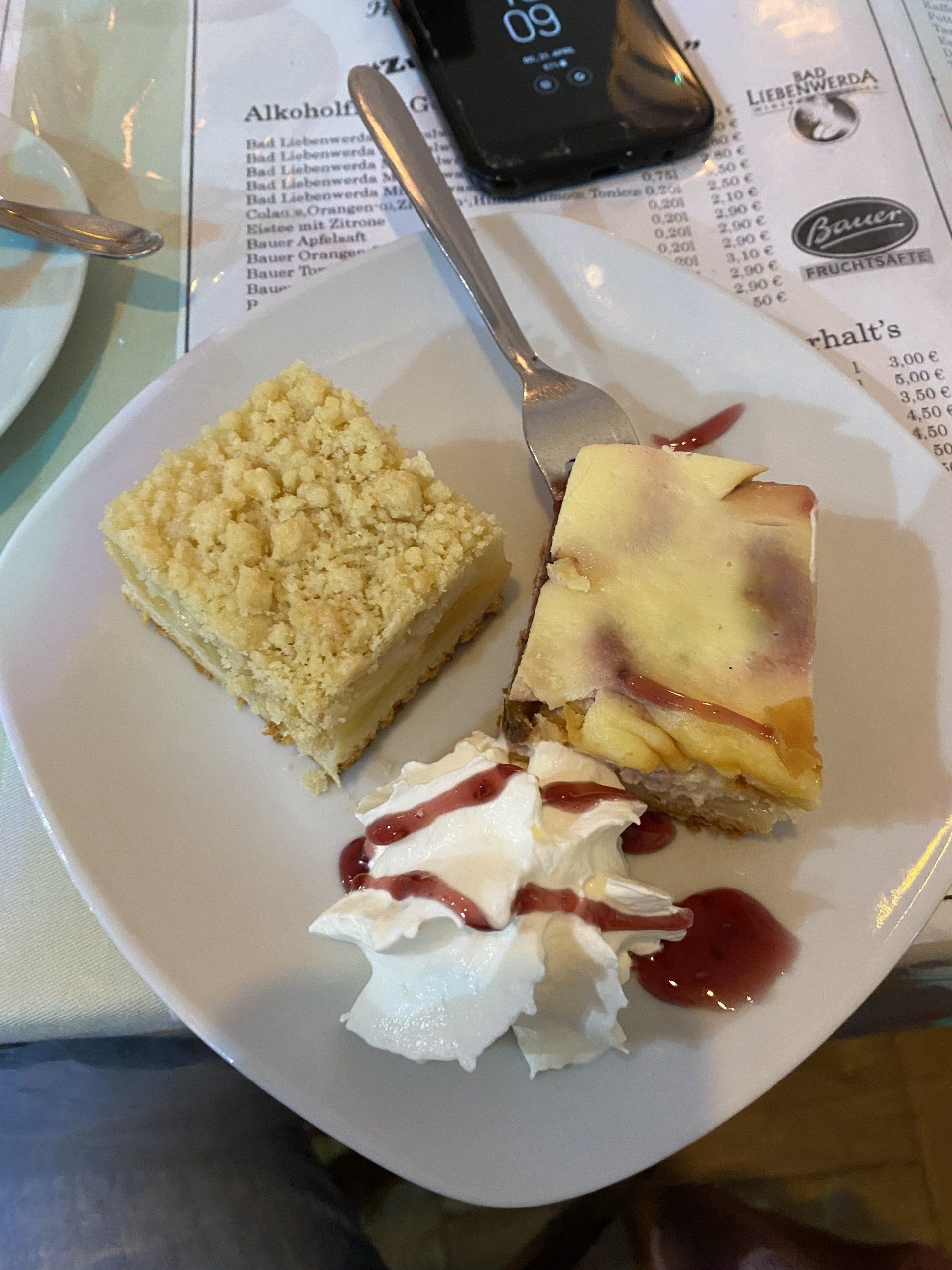 Dessertteller mit zwei Kuchenstücken, Schlagsahne und Beerensauce. Telefon und Gabel auf dem Tisch.