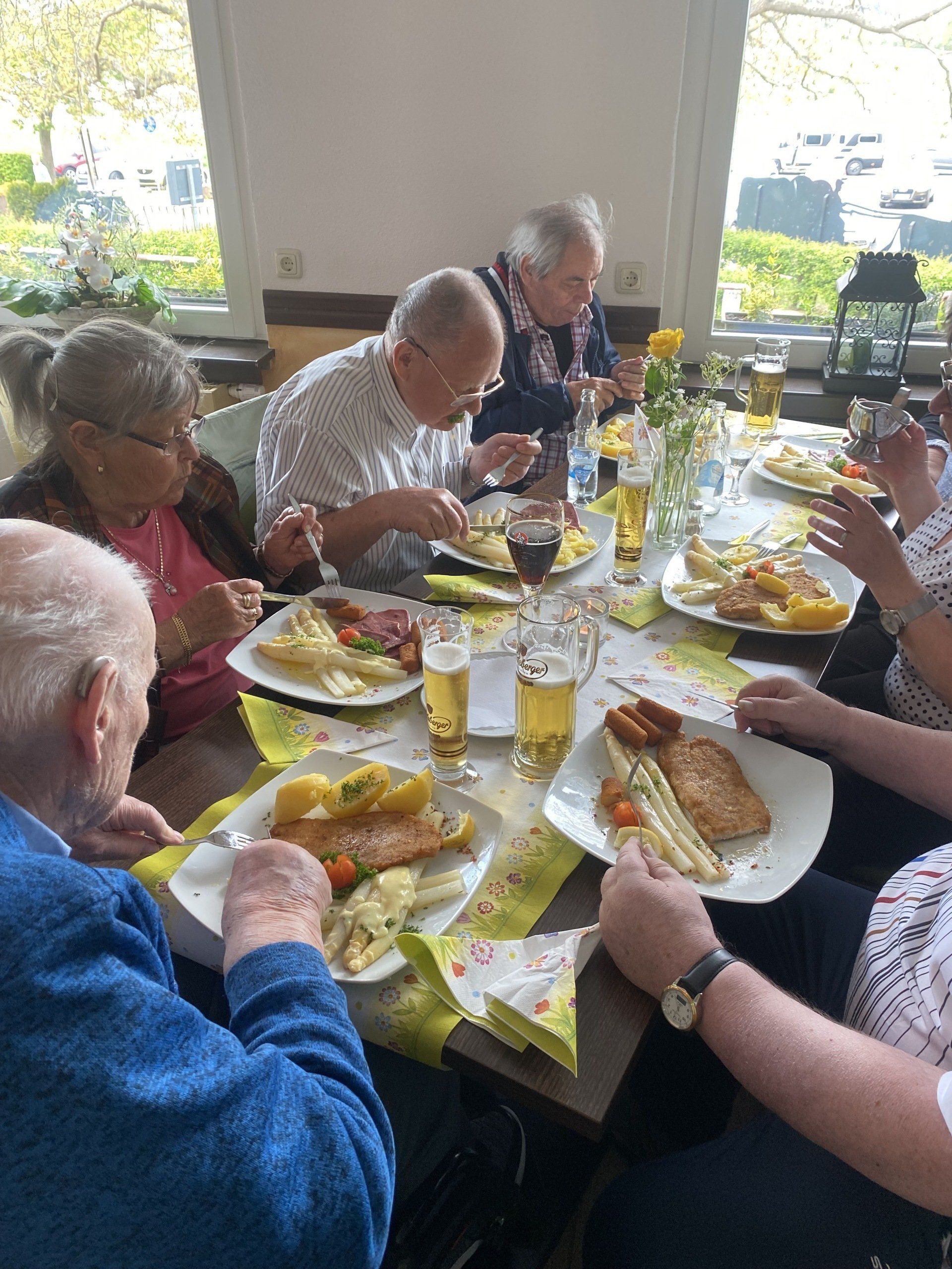 Menschen essen an einem Tisch. Teller mit Essen und Gläser sind sichtbar. Sonnenlicht fällt herein.