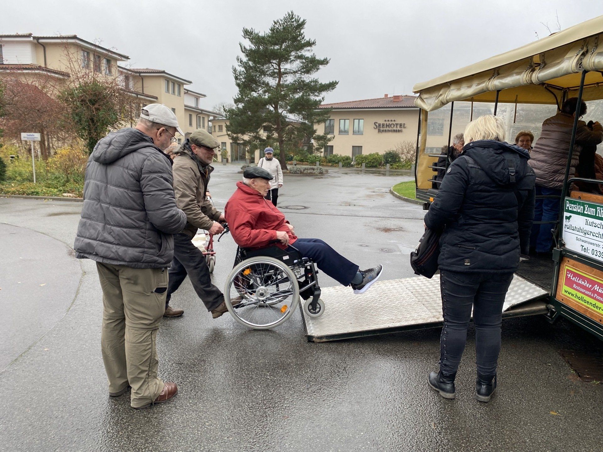 Menschen helfen einer Person im Rollstuhl die Rampe hinauf zu einem Imbisswagen. Bewölkter Tag.