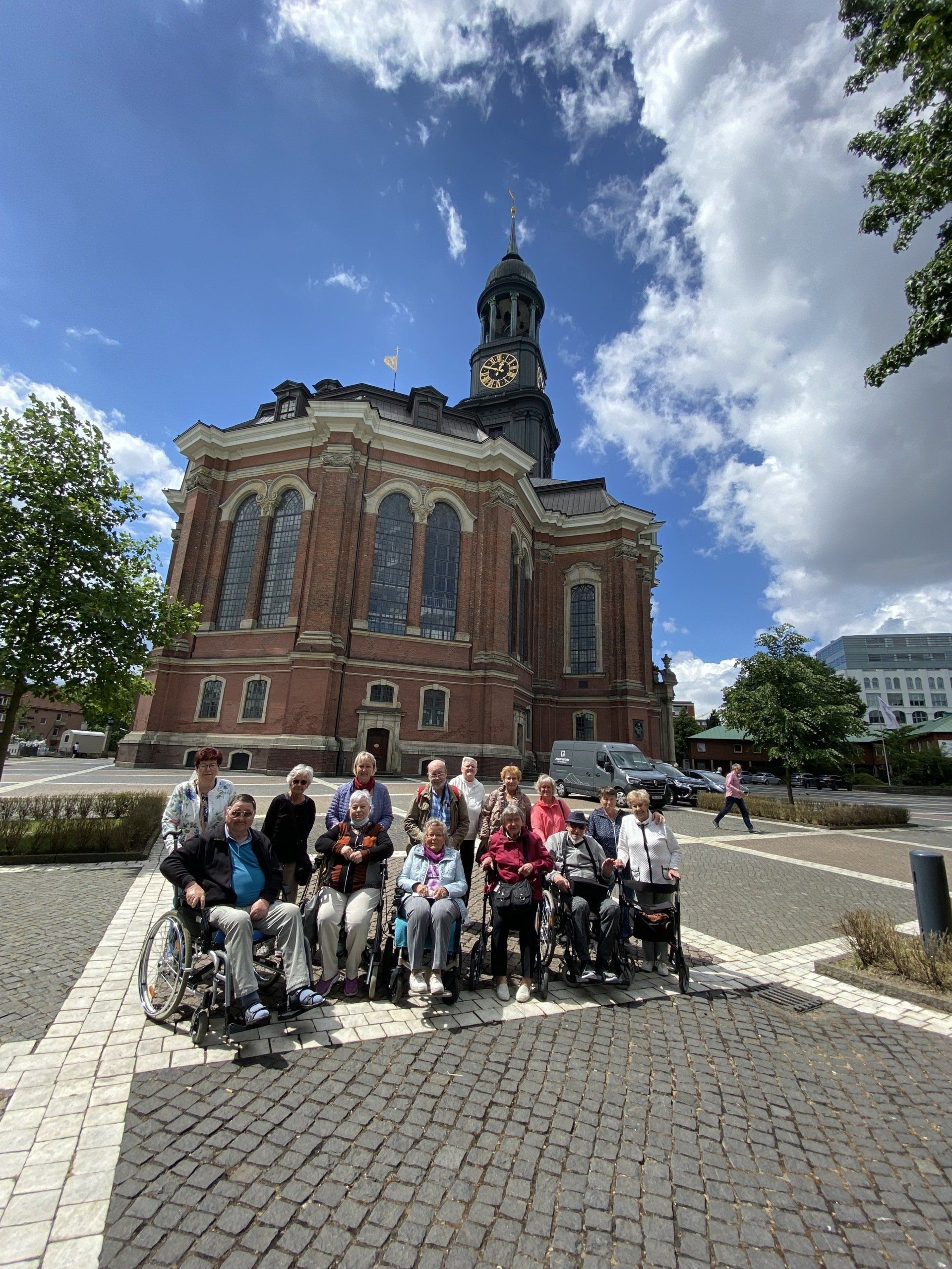 Gruppe in Rollstühlen vor einer Backsteinkirche mit hohem Kirchturm an einem sonnigen Tag.