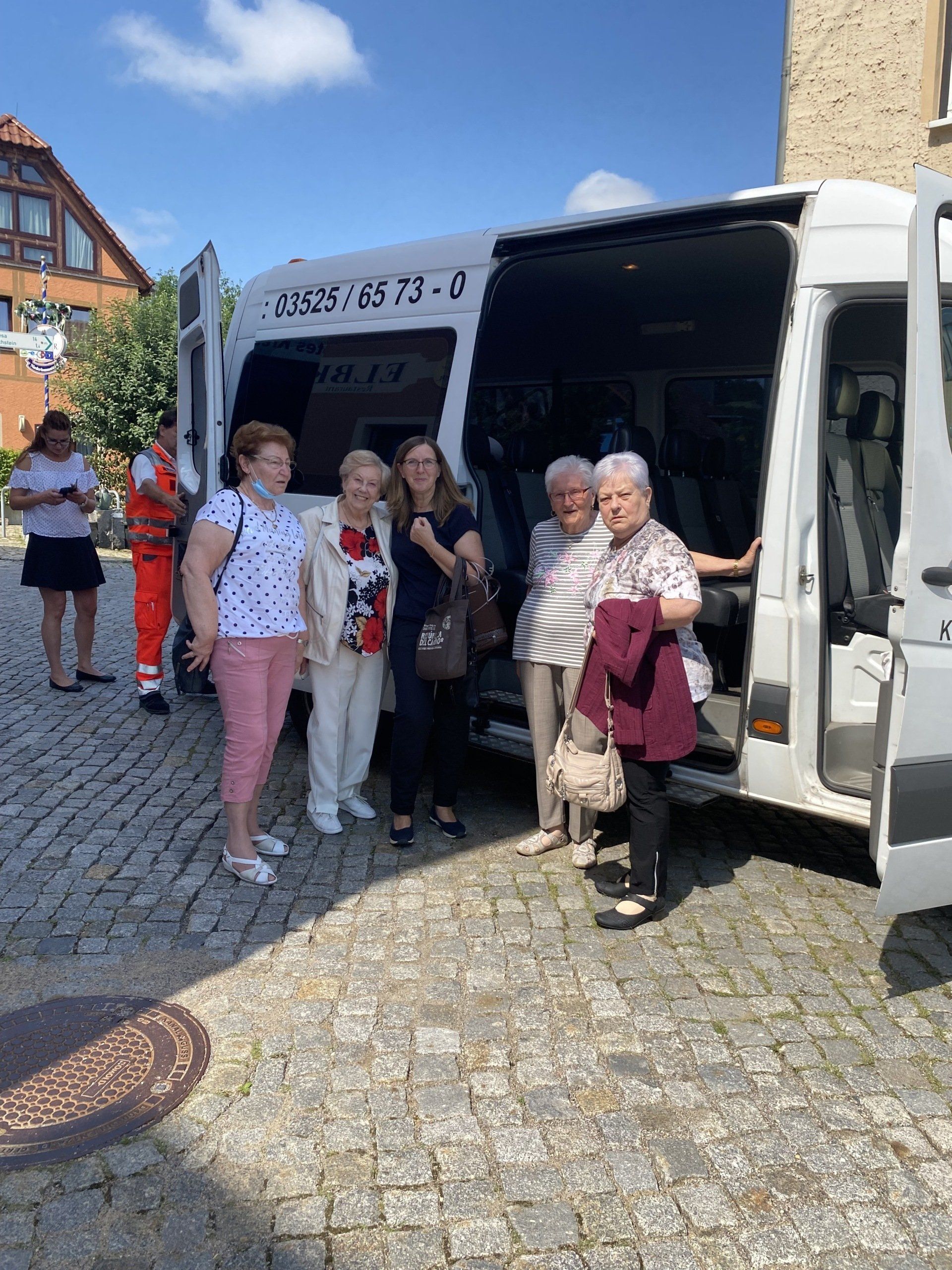 Eine Gruppe von Menschen steigt auf einer Kopfsteinpflasterstraße aus einem weißen Lieferwagen aus; sonniger Tag.