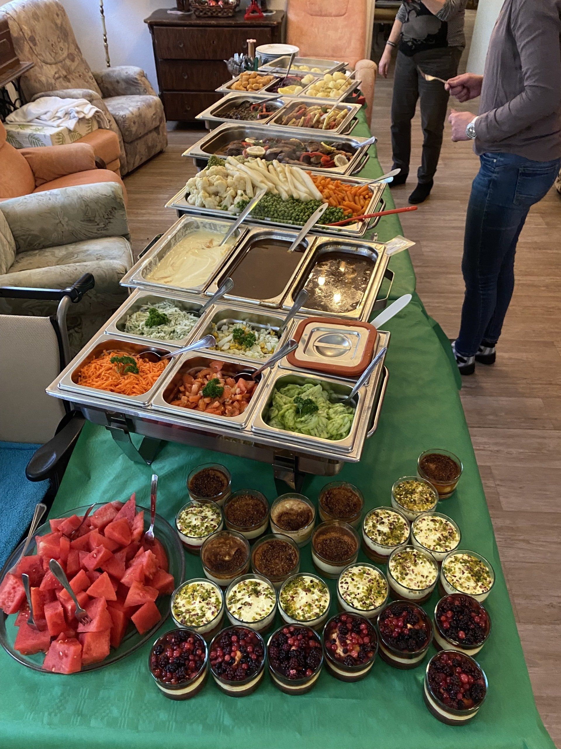 Buffettisch mit verschiedenen Gerichten; Wassermelone, Salate, Fleisch, Desserts. Leute stehen in der Nähe.