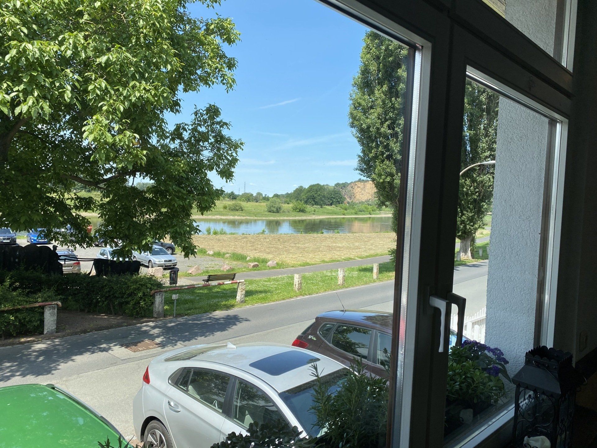 Blick durch ein Fenster auf eine Straße, Autos, Bäume und einen See unter blauem Himmel.