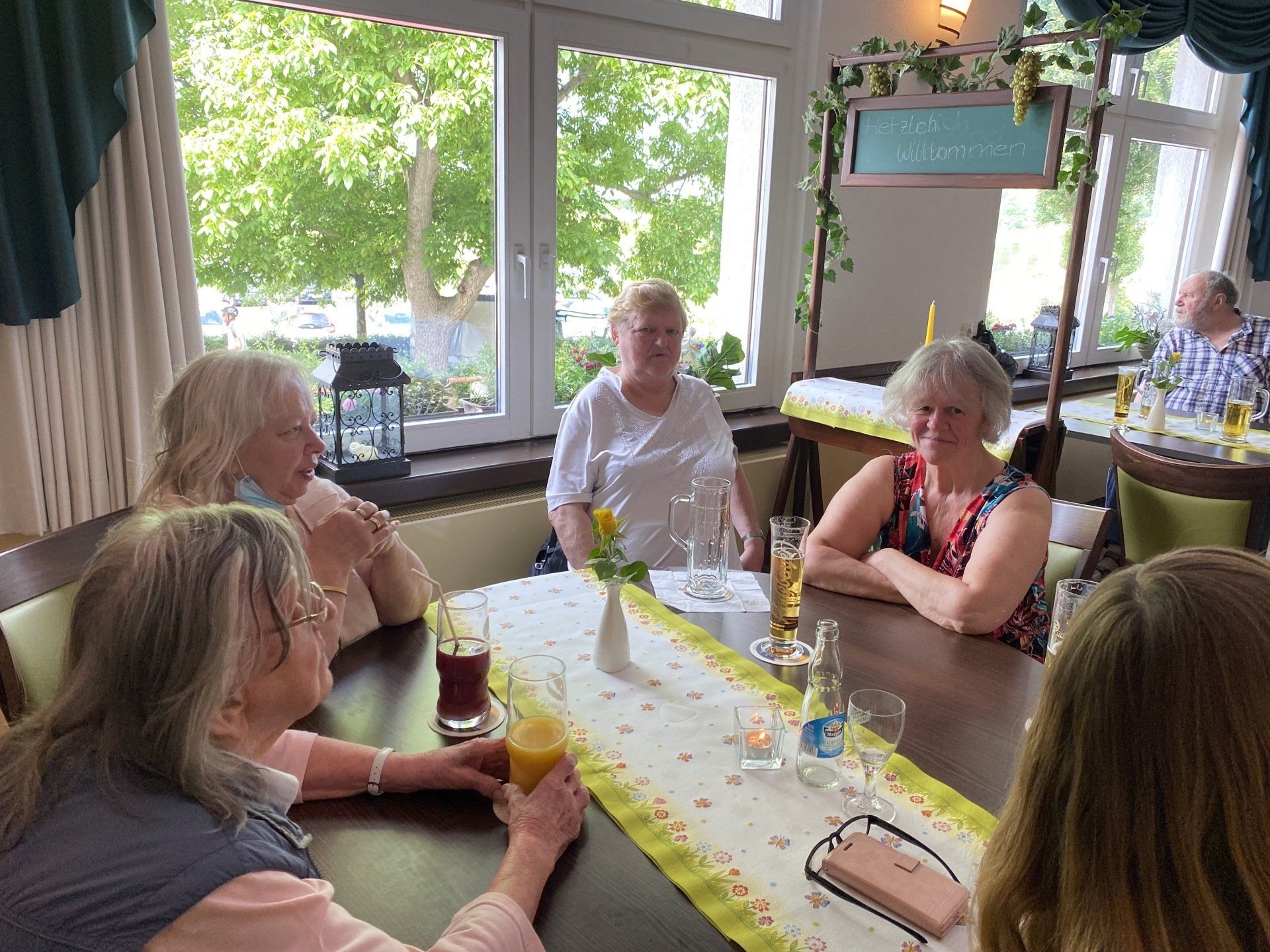 Menschen sitzen an einem Tisch in einem Restaurant. Sonnenlicht fällt durch große Fenster. Einige blicken in die Kamera.
