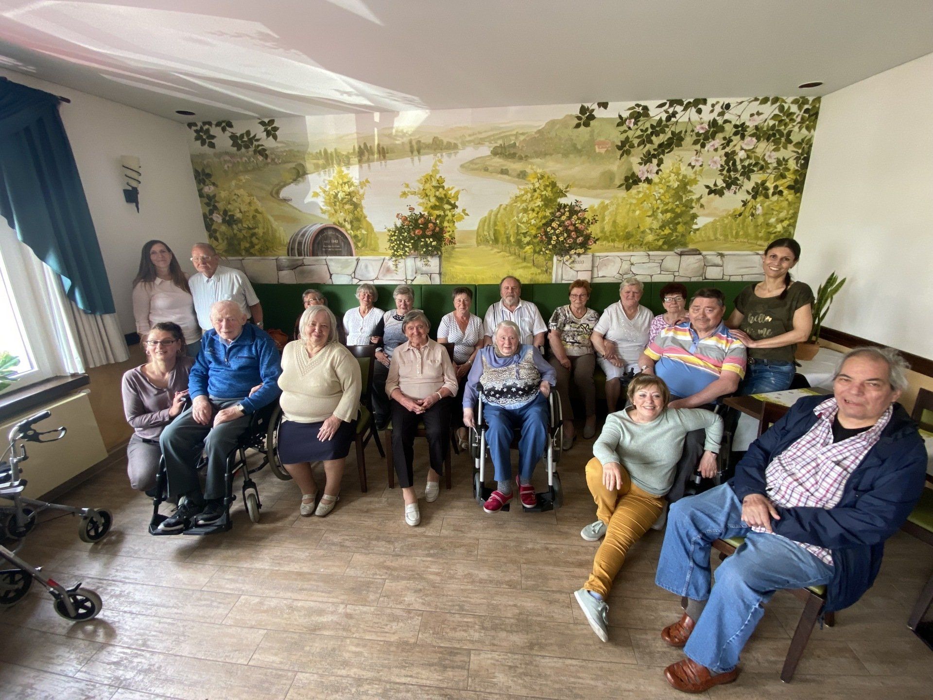 Gruppe von Menschen in einem Raum, einige davon im Rollstuhl. Landschaftsgemälde an der Wand.