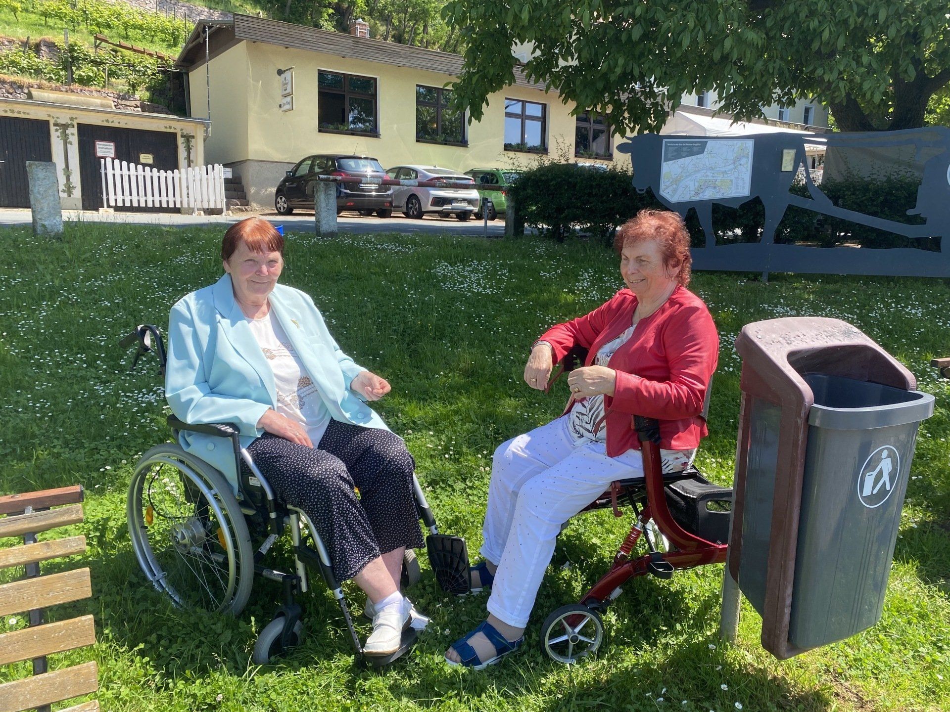 Zwei Frauen, eine im Rollstuhl, die andere mit einem Rollator, sitzen im Freien neben einer Mülltonne.