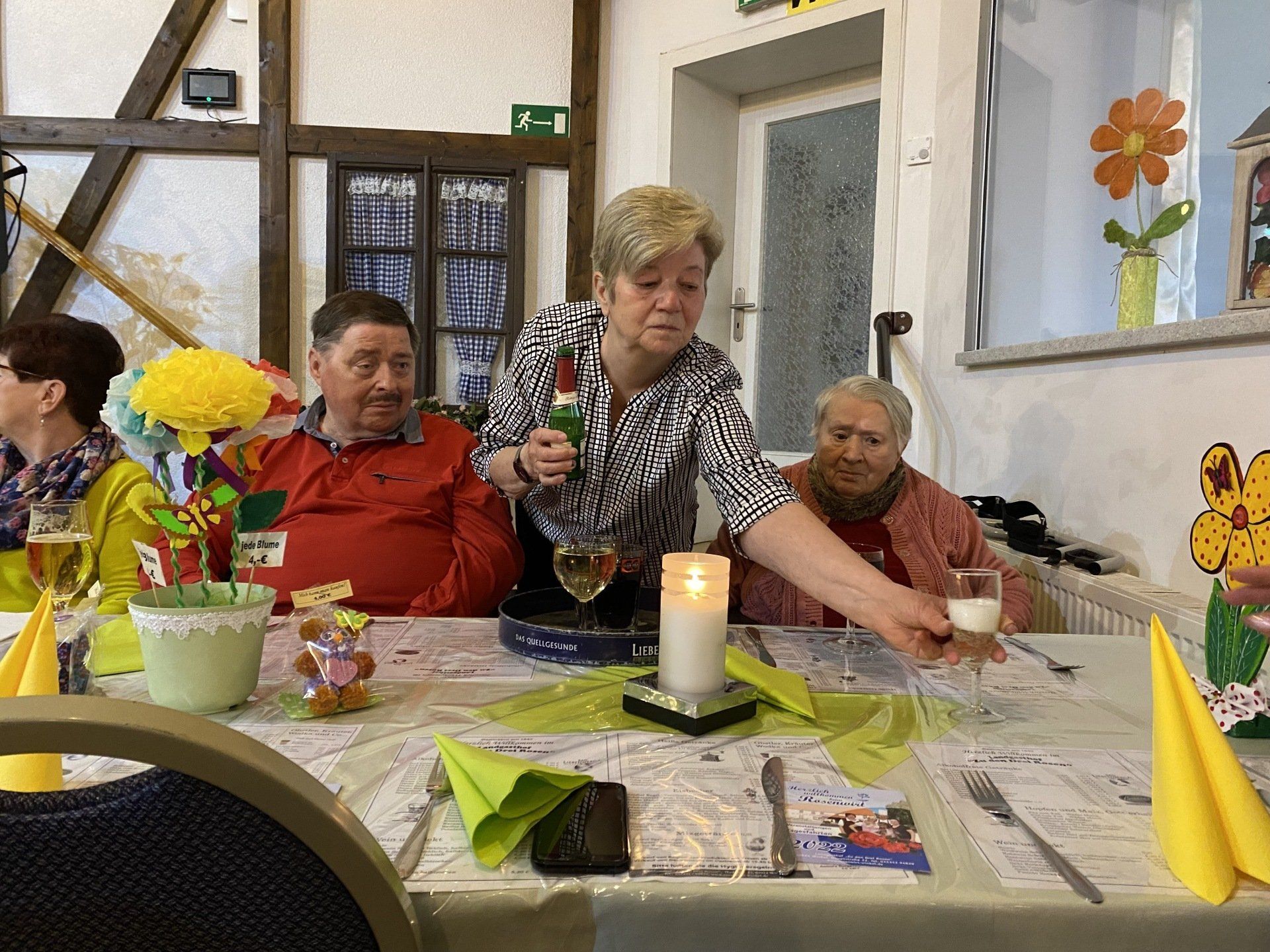 Eine Frau schenkt an einem festlich gedeckten Tisch mit drei sitzenden Personen, Blumen und Dekorationen ein Getränk ein.