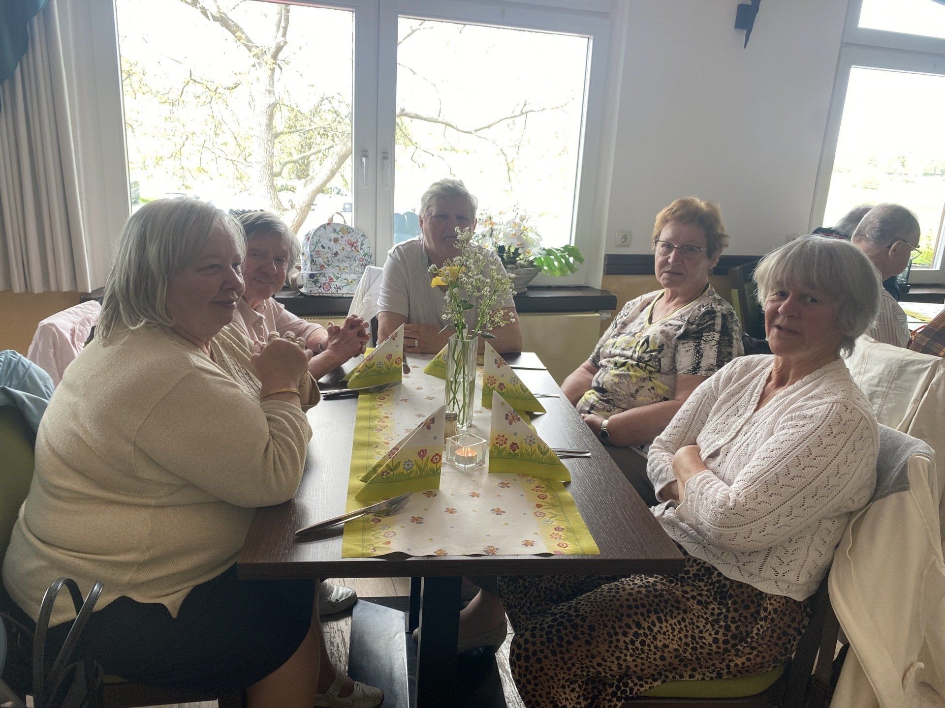Menschen sitzen an einem Tisch in einem Raum und unterhalten sich. Blumen und Servietten schmücken den Tisch. 