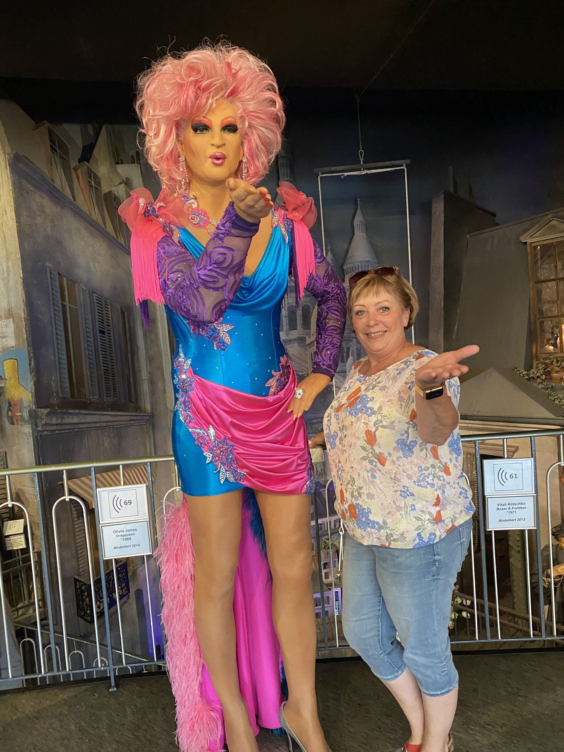 Frau posiert mit Dragqueen-Statue, beide werfen Küsse zu. Statue ist blau und rosa. Gebäudehintergrund.