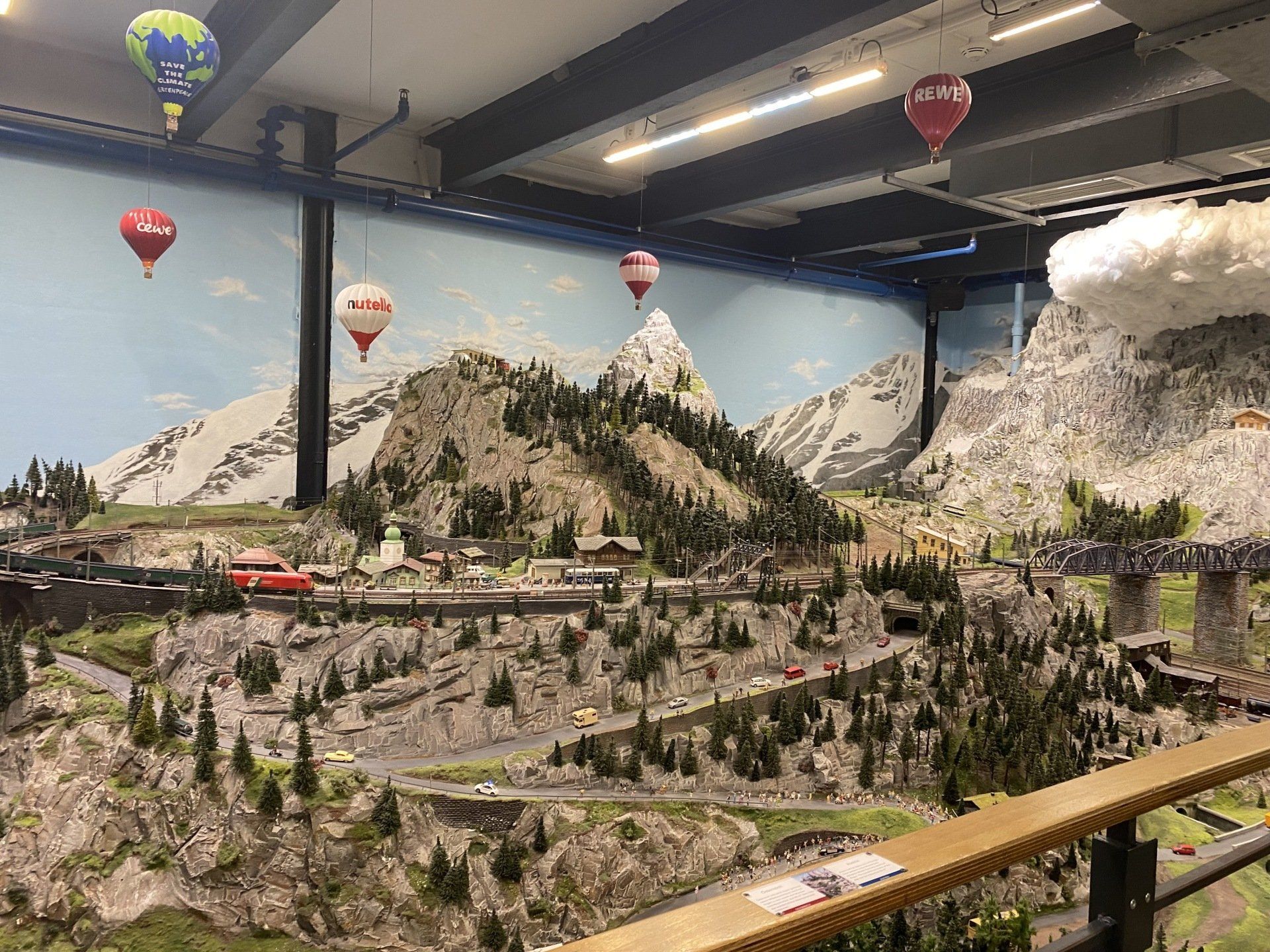 Miniaturlandschaft mit Bergen, Bäumen, Straßen und Zügen. Bunte Heißluftballons hängen über ihnen.