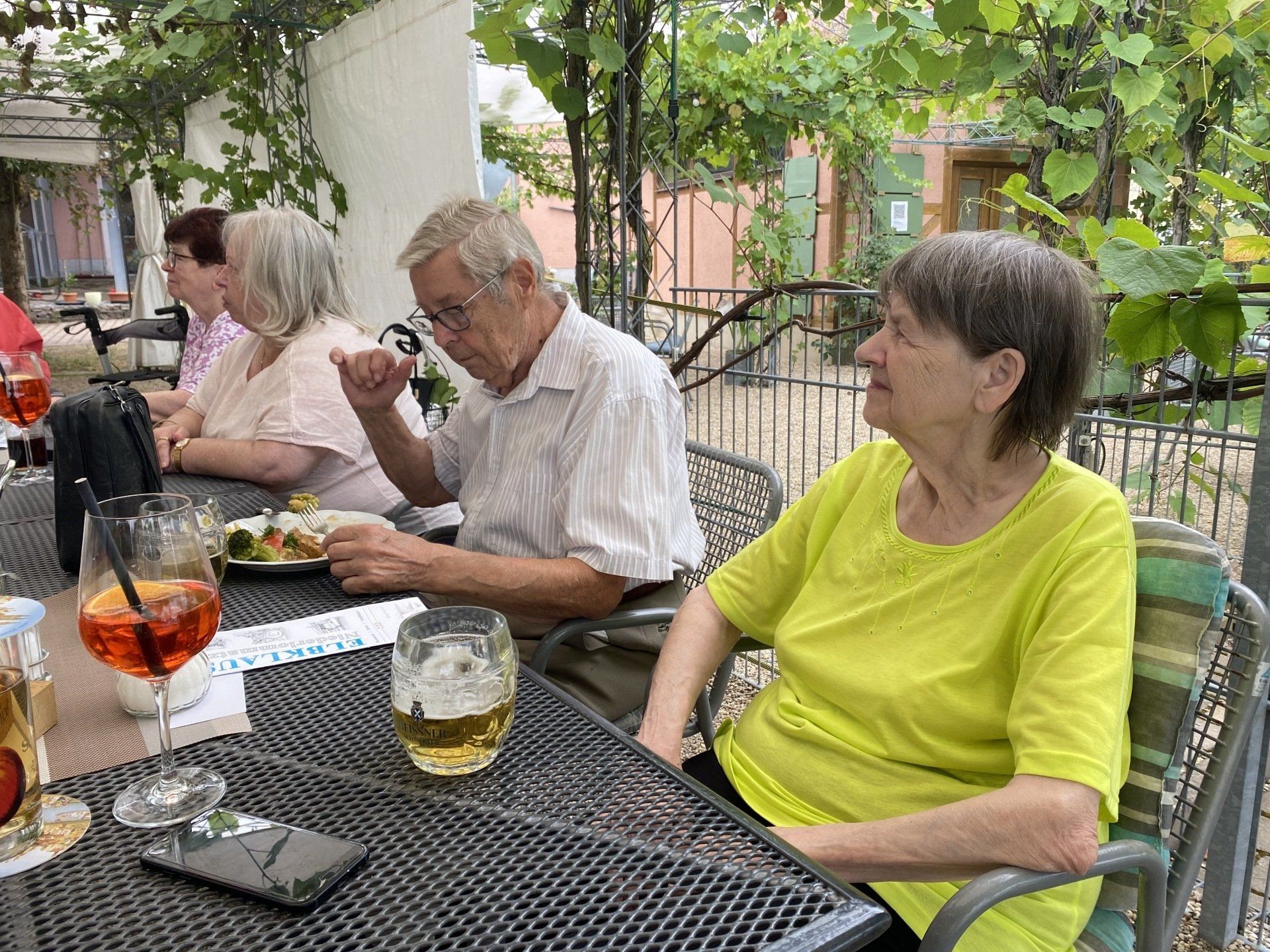 Menschen sitzen an Tischen im Freien, essen und trinken. Weinreben spenden Schatten.