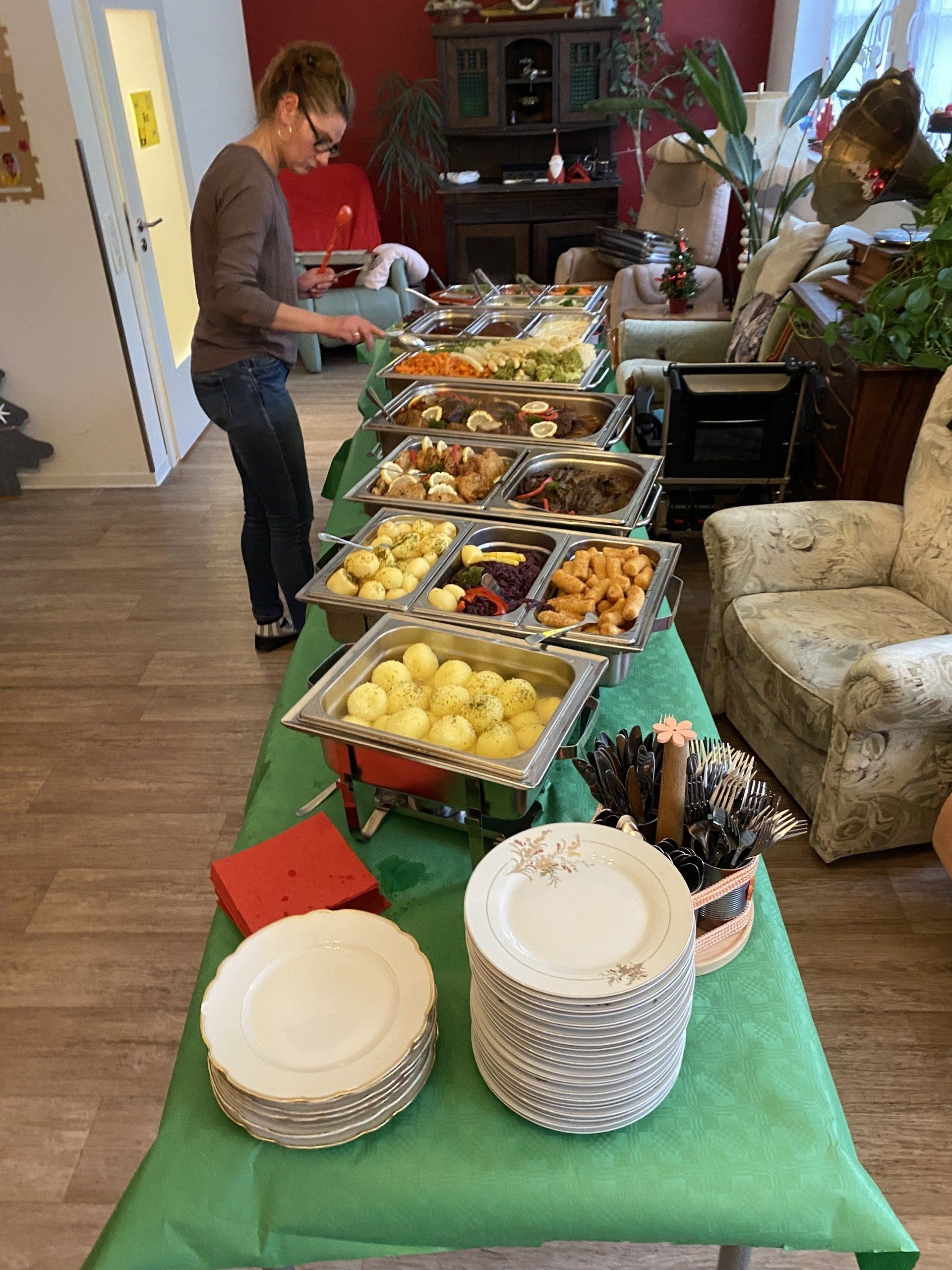 Buffettisch mit verschiedenen Gerichten, Person serviert Essen. Gestapelte Teller. Grüne Tischdecke in einem Raum.