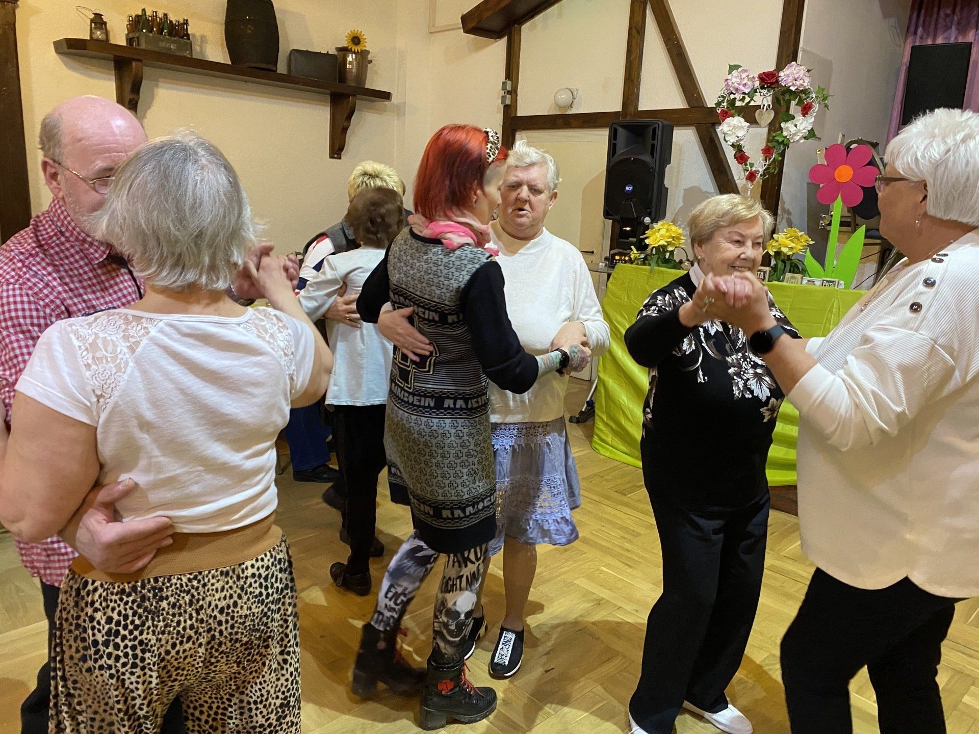 Menschen tanzen in einem Raum mit Holzbalken. Paare liegen in der Umarmung und amüsieren sich. Blumen schmücken den Raum.