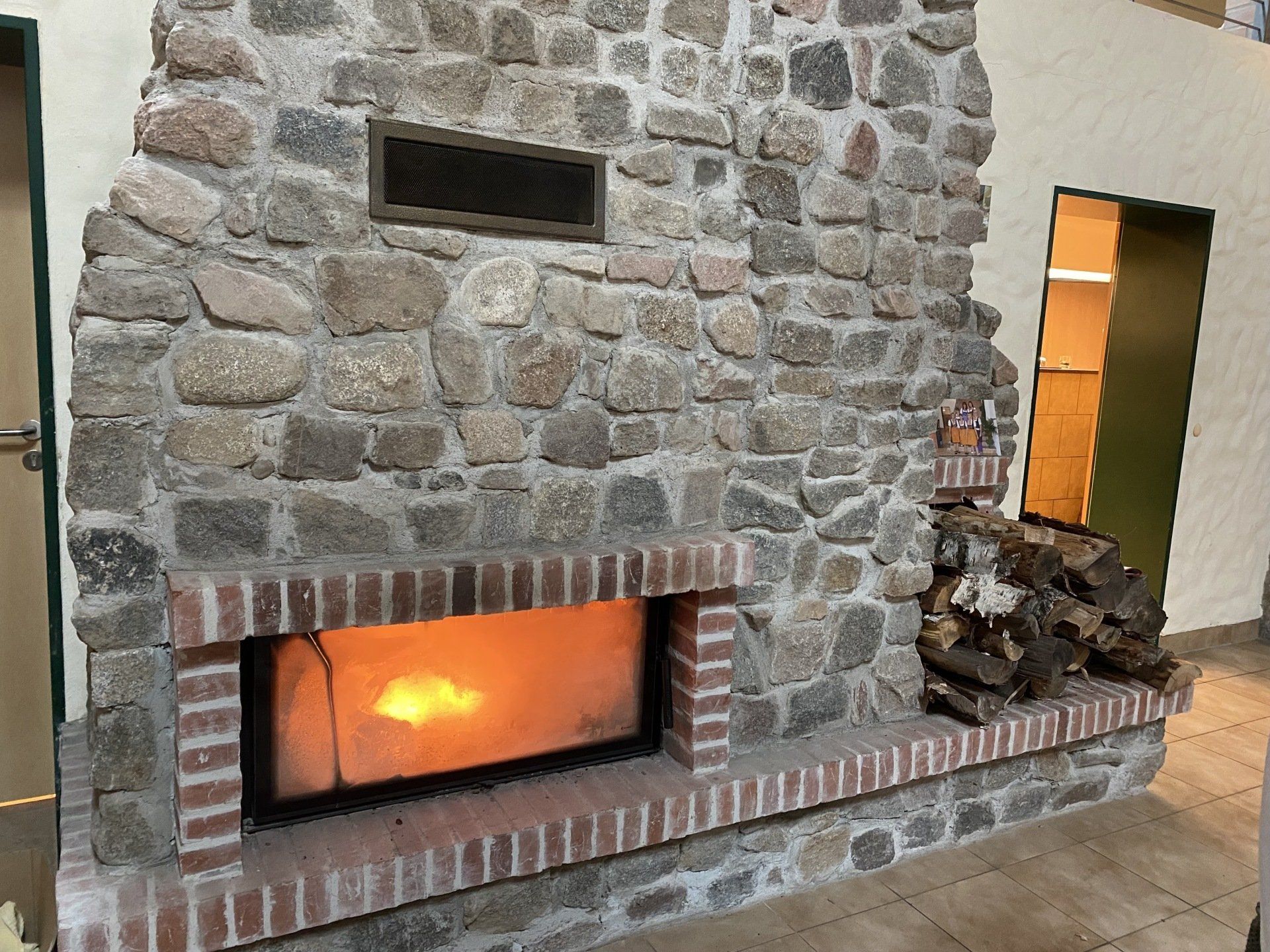 Kamin mit gemauerter Feuerstelle und Steinumrandung, Feuer brennt. Holzstapel an der Seite.