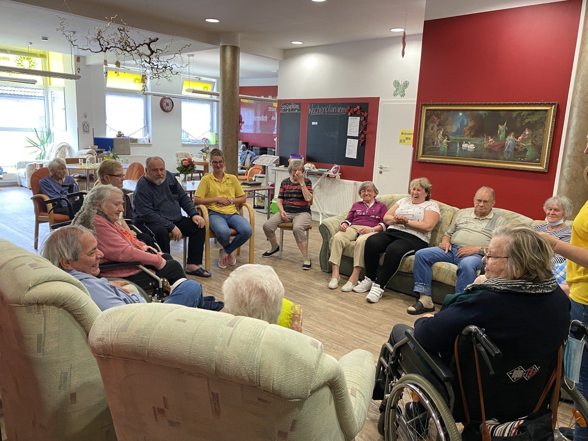 Eine Gruppe von Menschen sitzt im Kreis auf Stühlen und Sofas in einem Gemeinschaftsraum mit roter Wand.