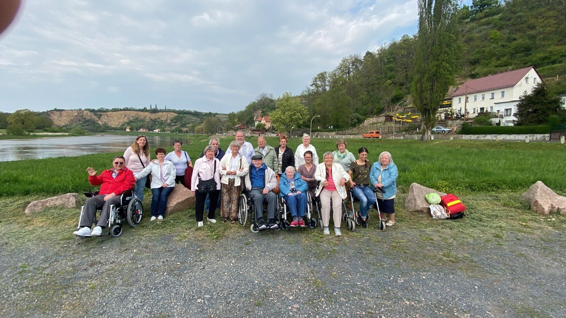 Eine Gruppe von Menschen, einige davon im Rollstuhl, steht an einem Gewässer und einem Hügel. Einige winken.
