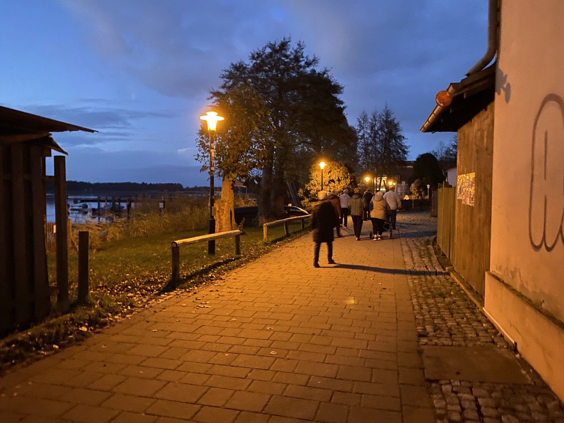 Kopfsteinpflasterweg, beleuchtet von Straßenlaternen, neben Wasser. Menschen gehen in der Dämmerung den Weg entlang.