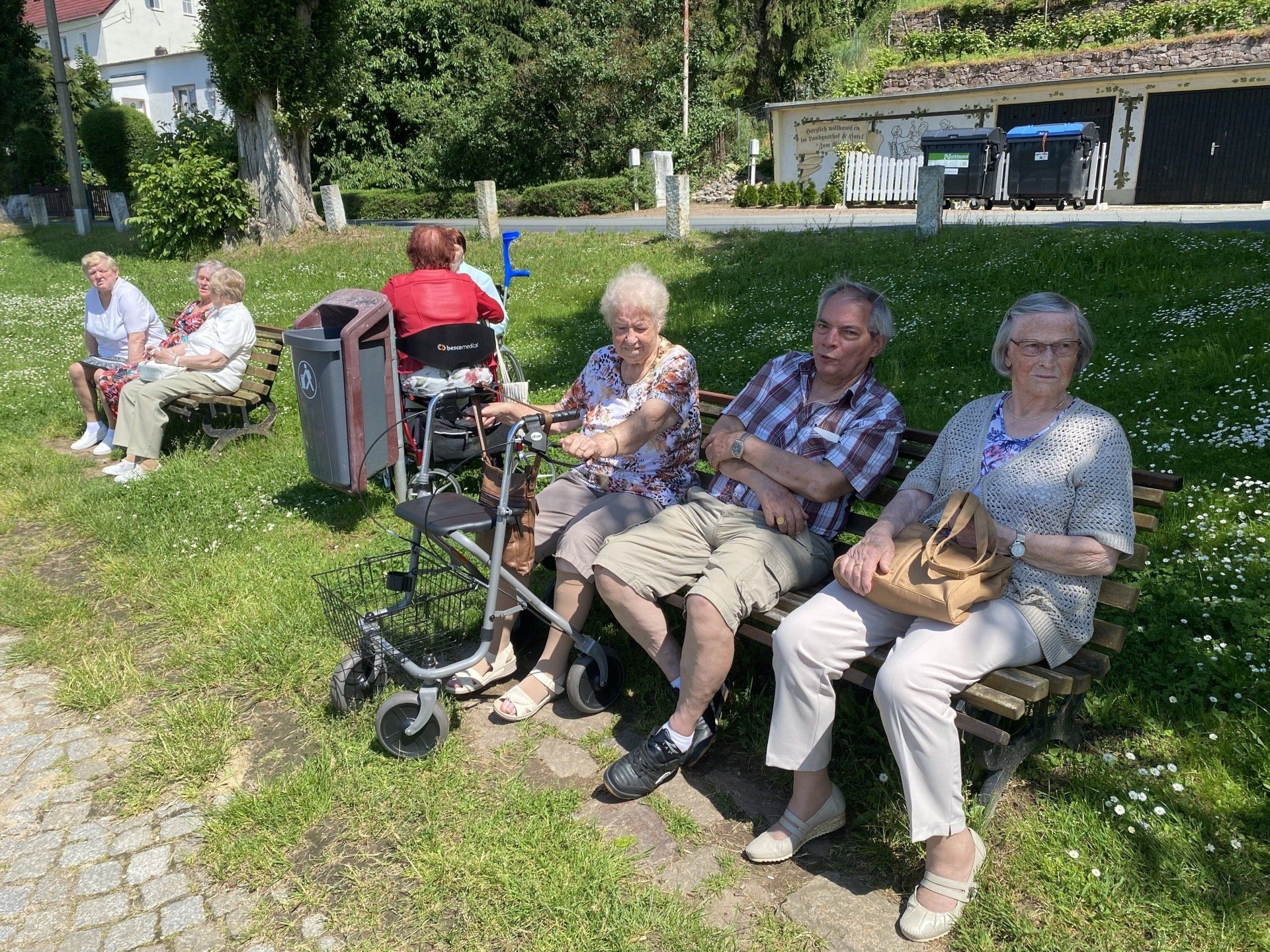 Eine Gruppe von Menschen sitzt an einem sonnigen Tag im Freien auf Parkbänken, einige davon mit Gehhilfen.