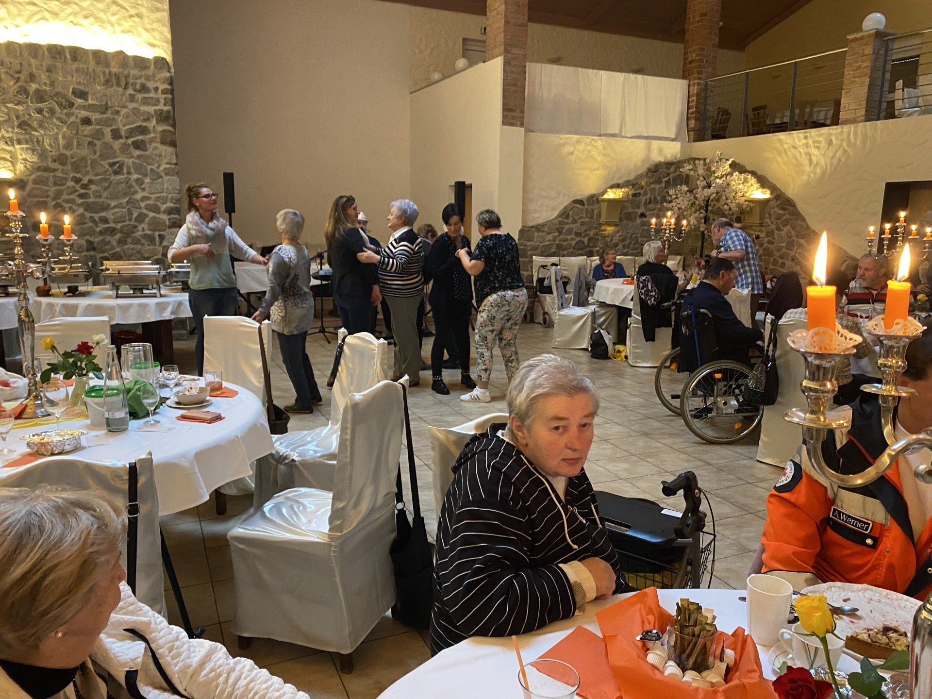 Menschen an Tischen, einige in Rollstühlen, in einem Saal mit Steinwänden, Kerzen und einem Buffet.