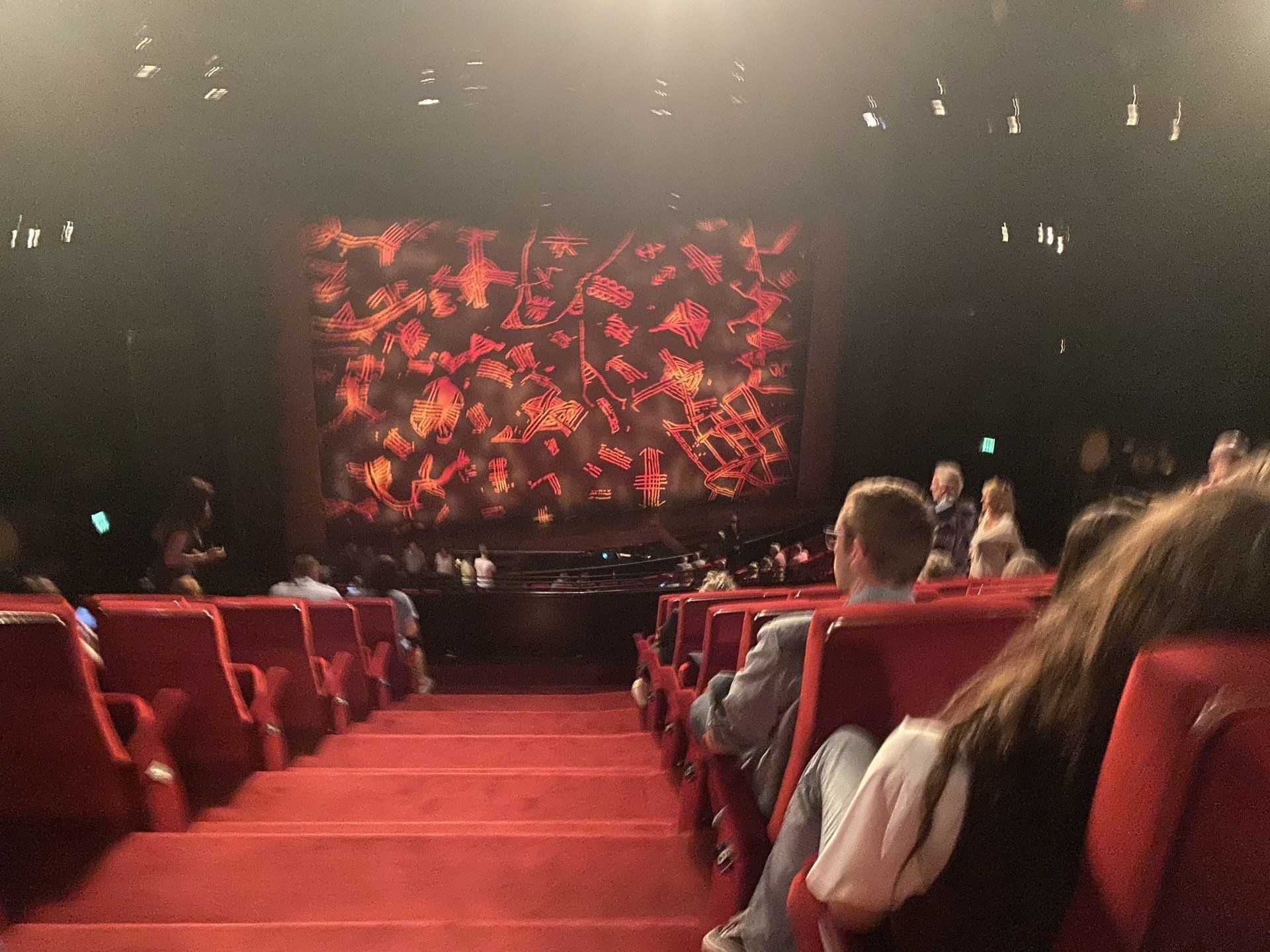 Publikum in einem Theater, rote Sitze vor einer beleuchteten Bühne mit abstrakten roten und orangefarbenen Bildern.
