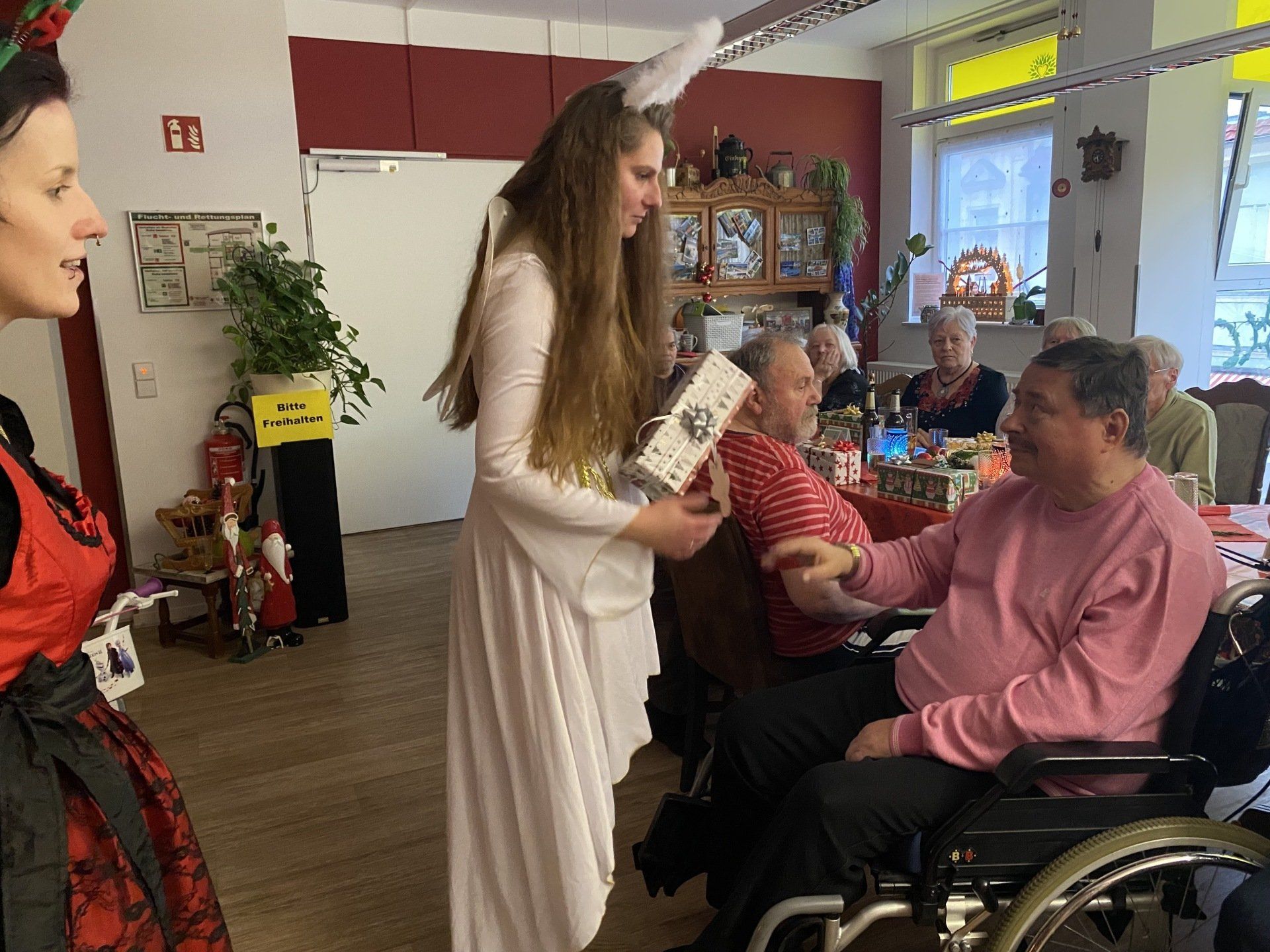 Frau mit Hasenohren bietet Mann im Rollstuhl ein Geschenk an; andere Leute an Tischen.