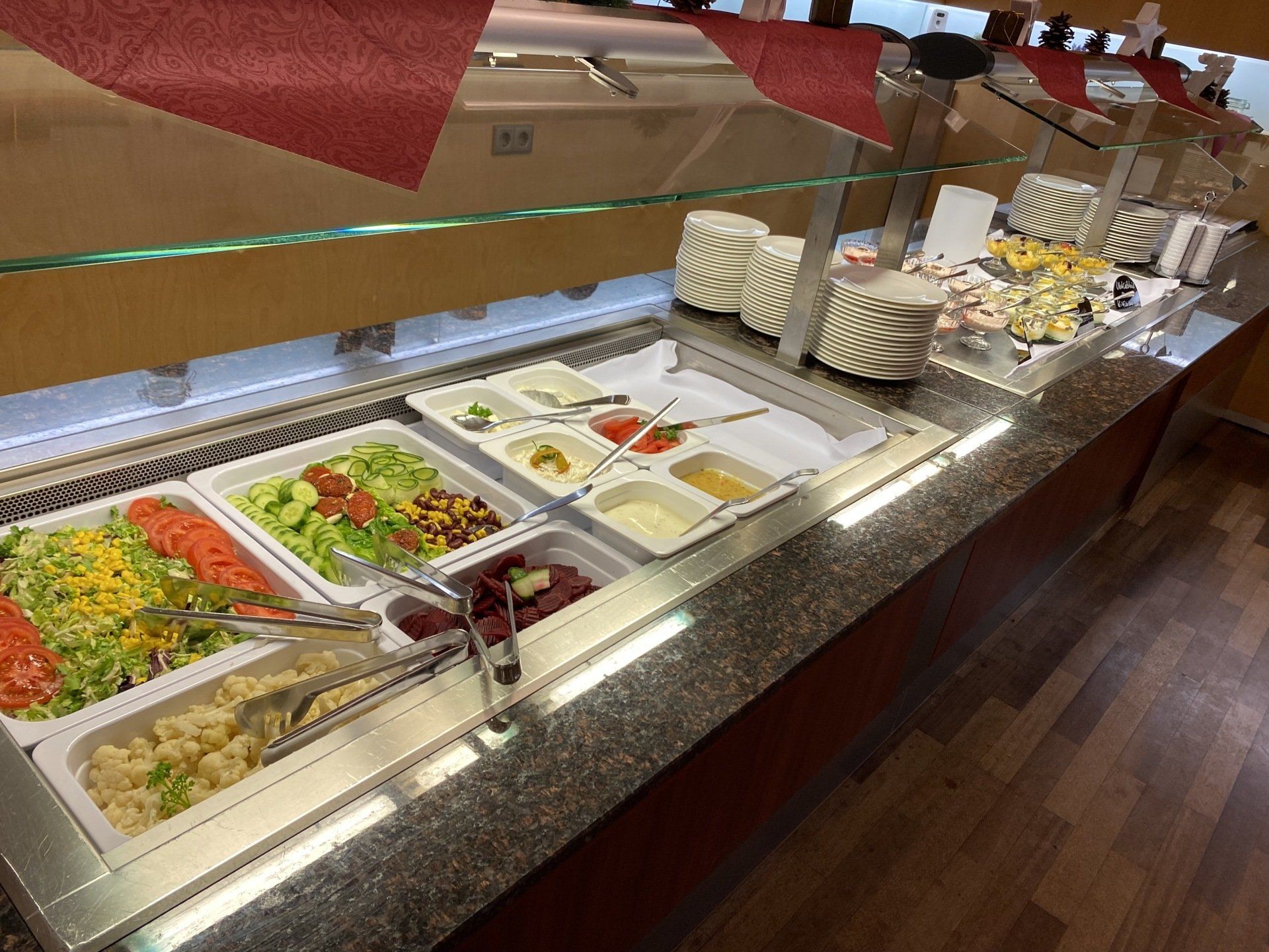Salatbar mit verschiedenen Zutaten in weißen Behältern, mit roten Servietten und Holzboden.
