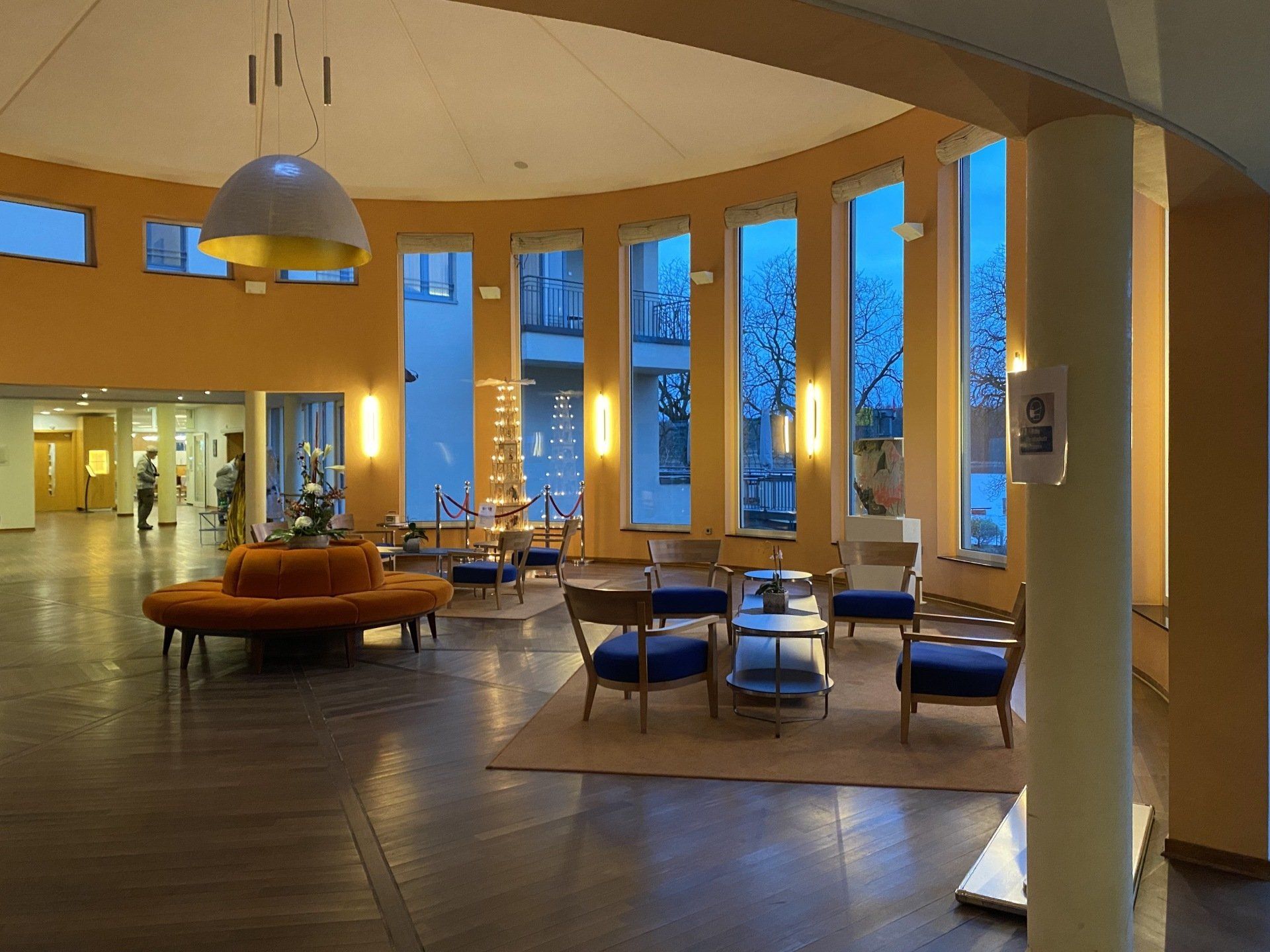 Hotellobby mit rundem Sitzbereich und großen Fenstern. Orange Wände, blaue Stühle, Holzboden.