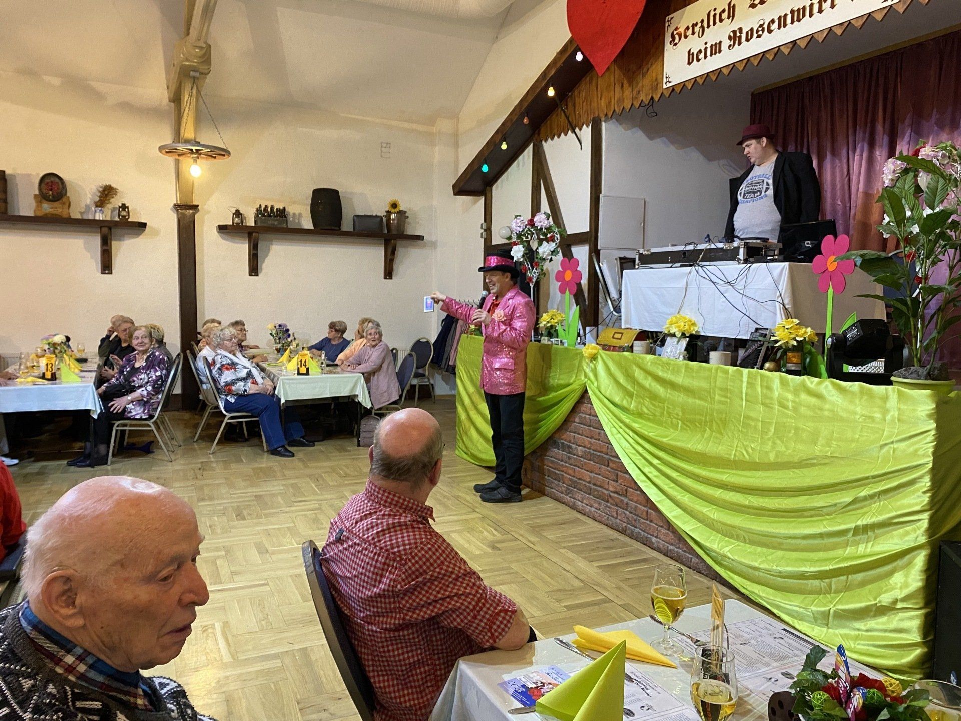 Mann in rosa Jacke tritt auf der Bühne auf; Publikum an Tischen im Saal.