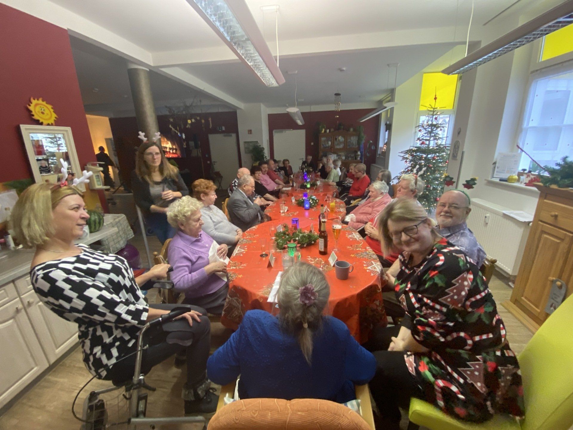 Eine Gruppe von Menschen sitzt an einem langen Tisch und feiert in einem weihnachtlich geschmückten Raum.