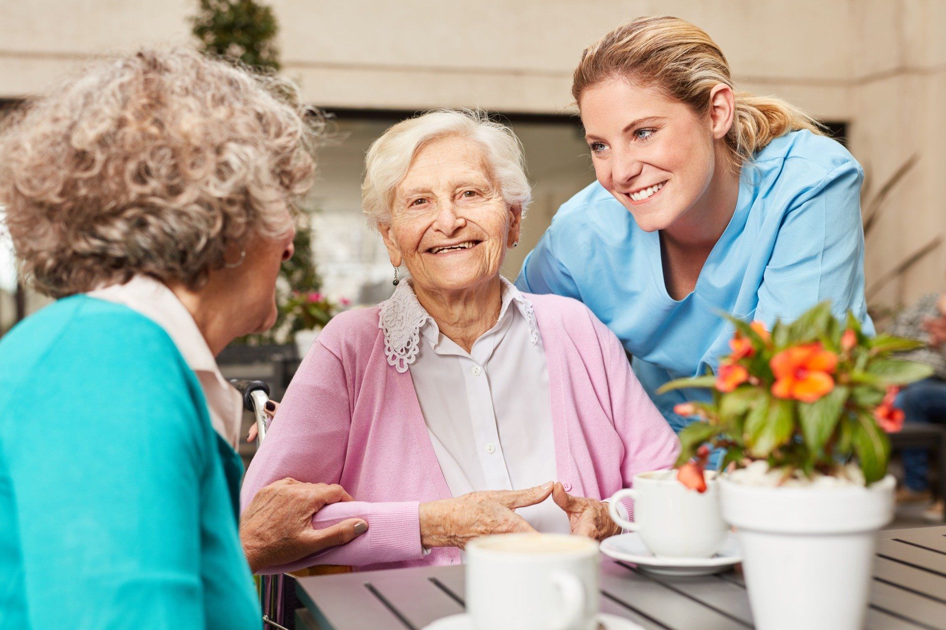 Zwei Frauen und eine Pflegekraft lächeln an einem Tisch im Freien mit Kaffeetassen und Blumen.