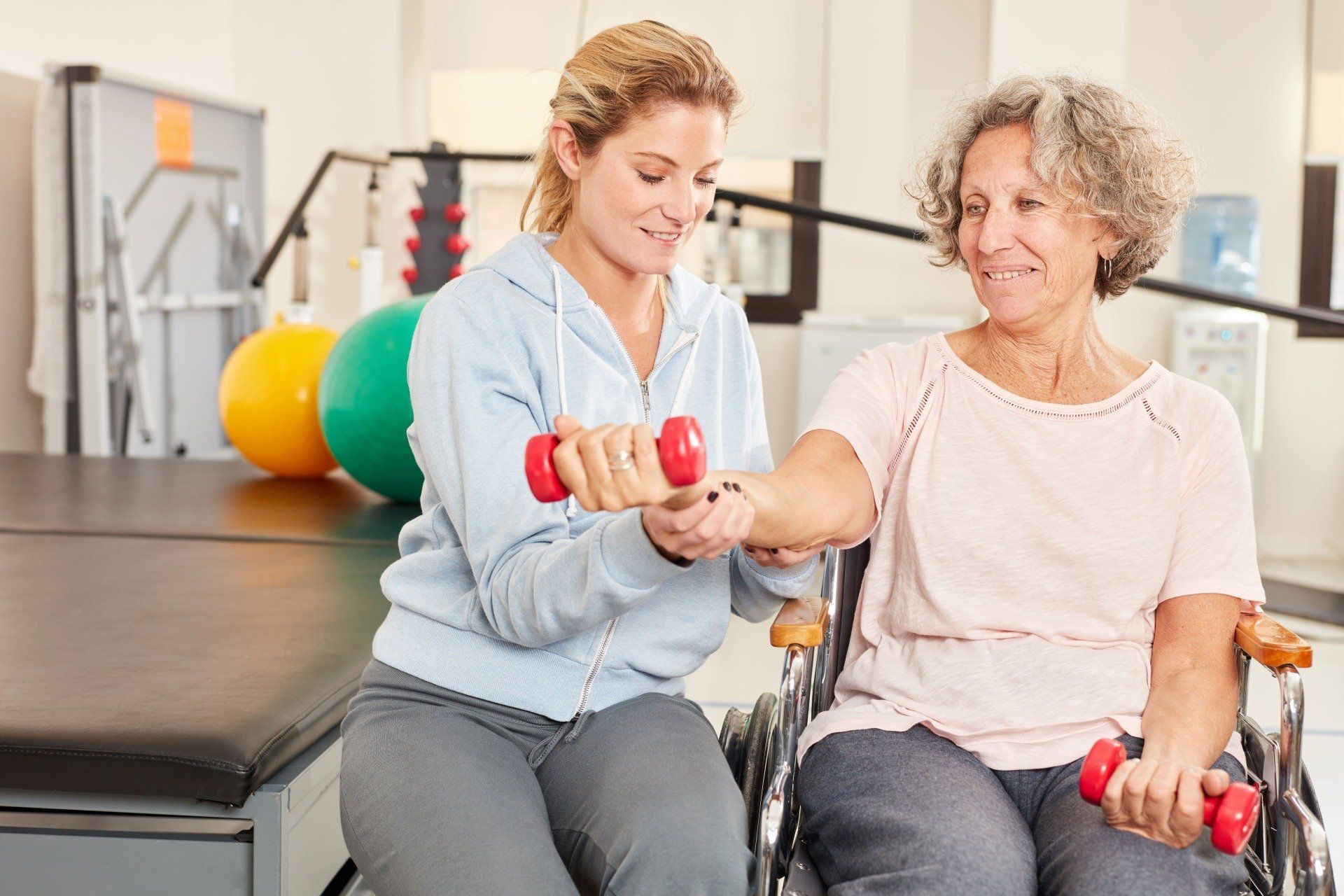 Eine Frau hilft einer Person im Rollstuhl bei Armübungen mit kleinen roten Hanteln in einem Therapiestudio.