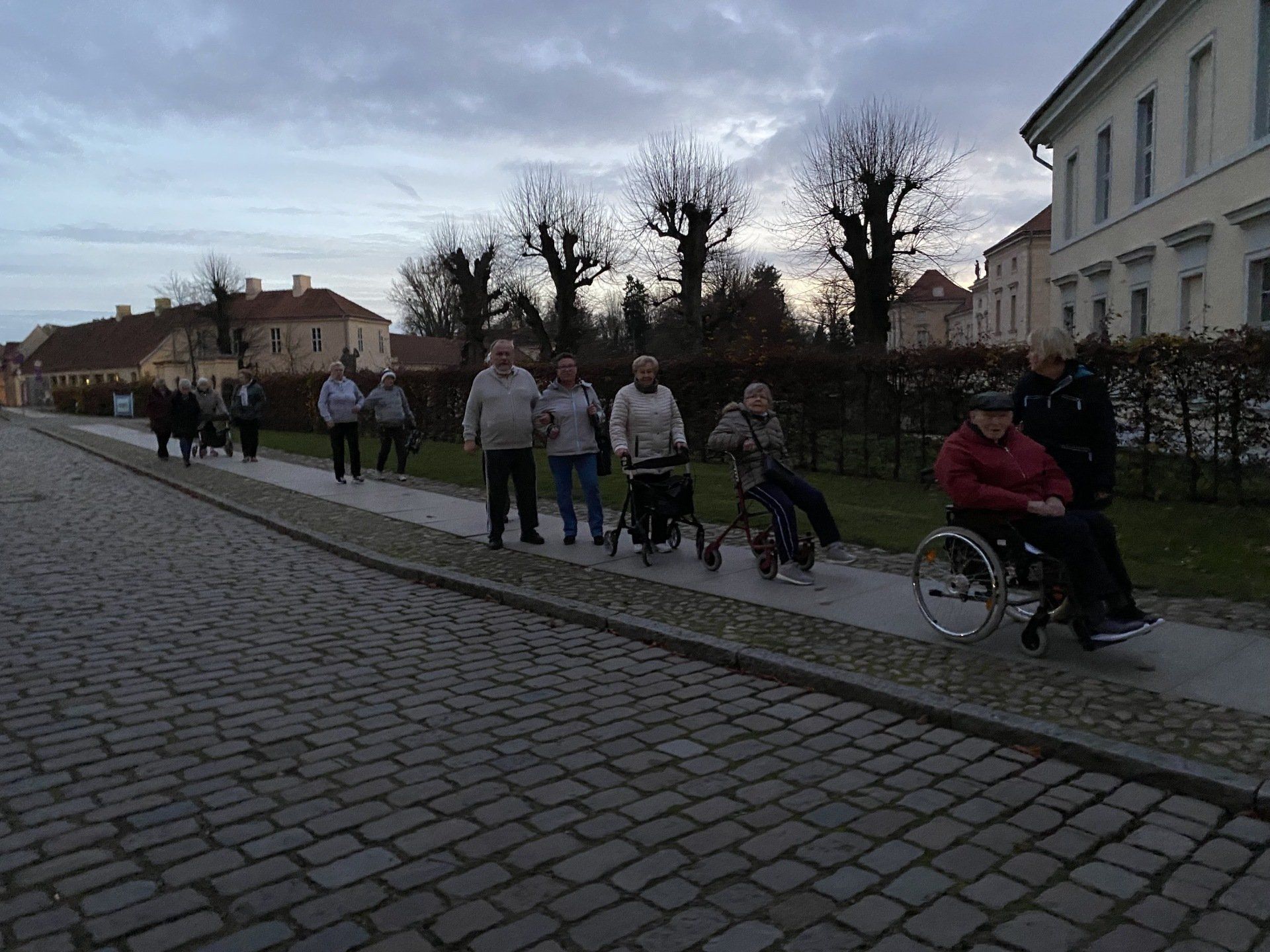 Eine Gruppe von Menschen, einige davon im Rollstuhl oder mit Gehhilfe, geht auf einem gepflasterten Weg vor einem Gebäude.