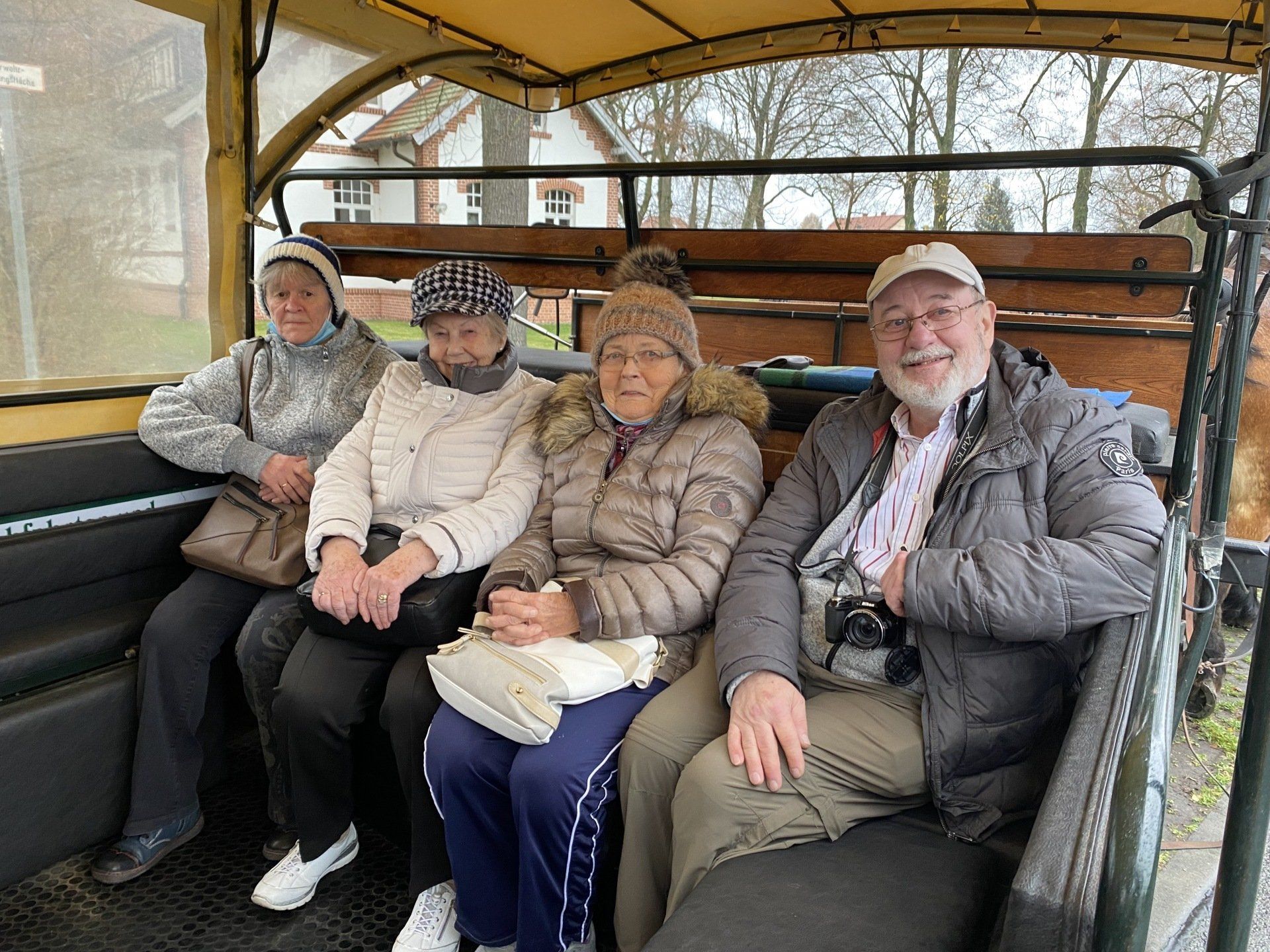 Vier Personen sitzen lächelnd in einem Planwagen. Der Wagen steht im Freien, im Hintergrund sind Bäume zu sehen.
