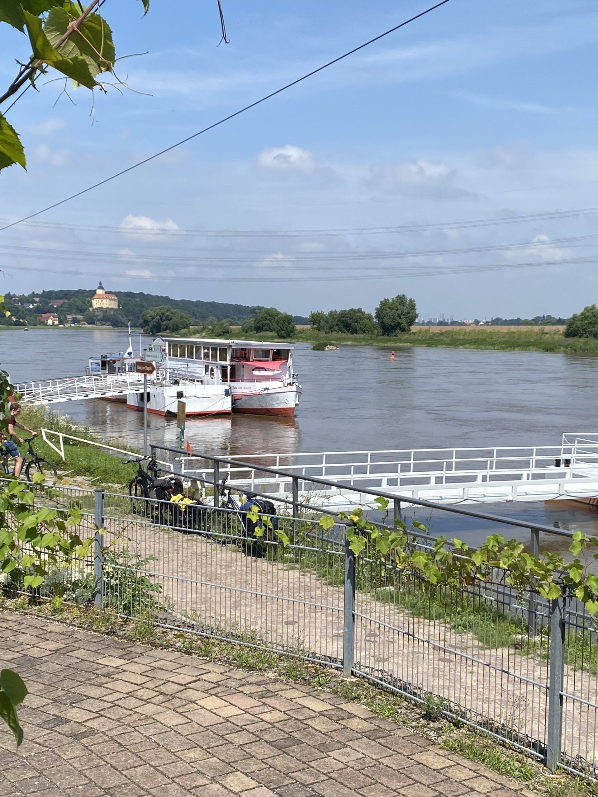 Boote liegen an einem Pier an einem Fluss. In der Ferne thront auf einem Hügel unter blauem Himmel eine Burg.