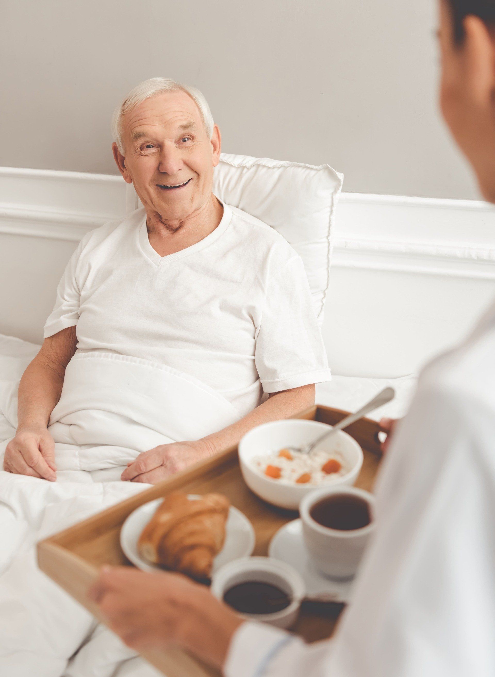 Mann im Bett lächelt Person mit Frühstückstablett an.