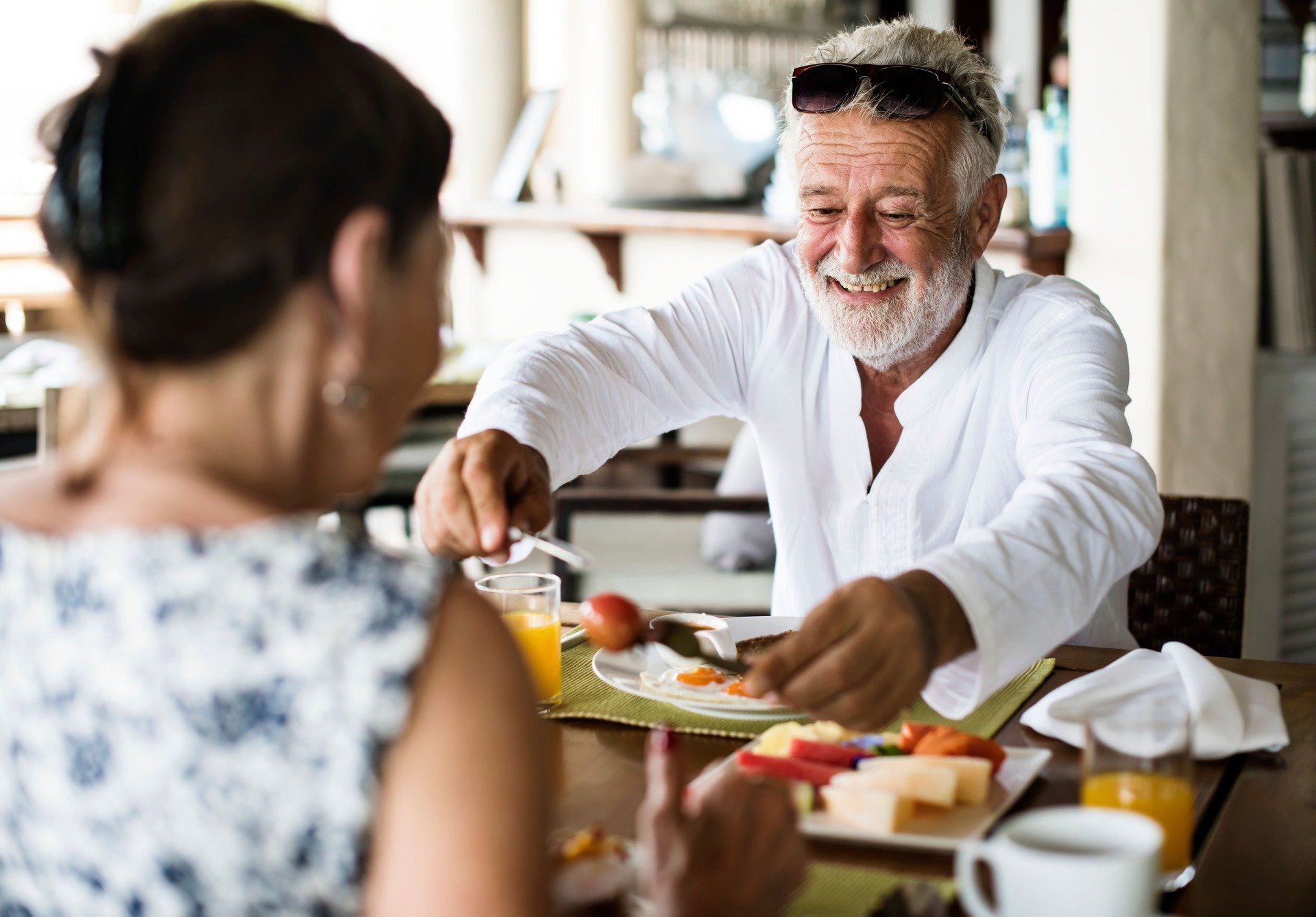 Lächelnder älterer Mann, der an einem Tisch mit einer Frau nach Essen greift. Essen und Getränke sind sichtbar. Heller Raum.