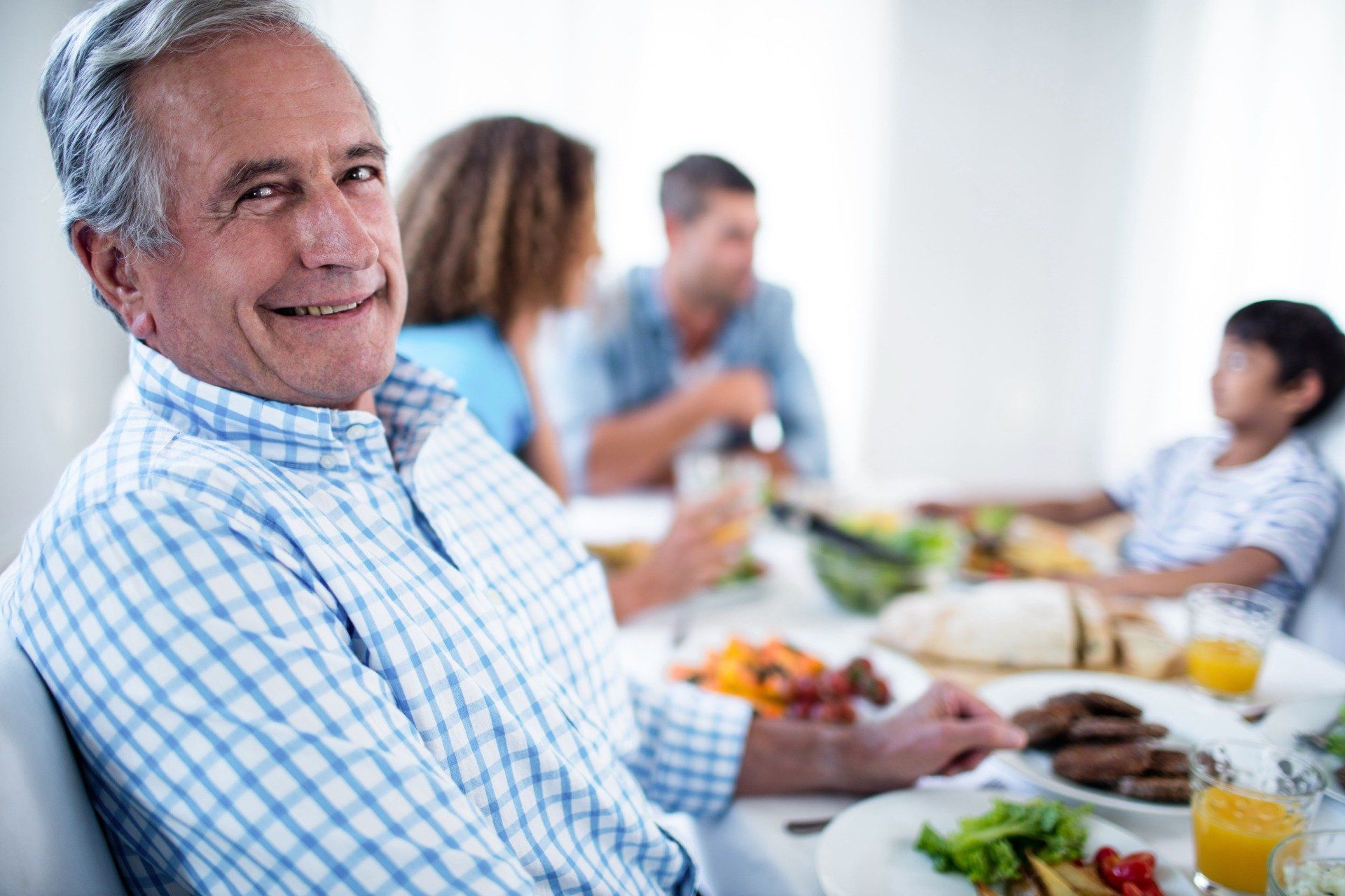 Senior lächelt in die Kamera, sitzt mit seiner Familie an einem Esstisch und genießt eine Mahlzeit.