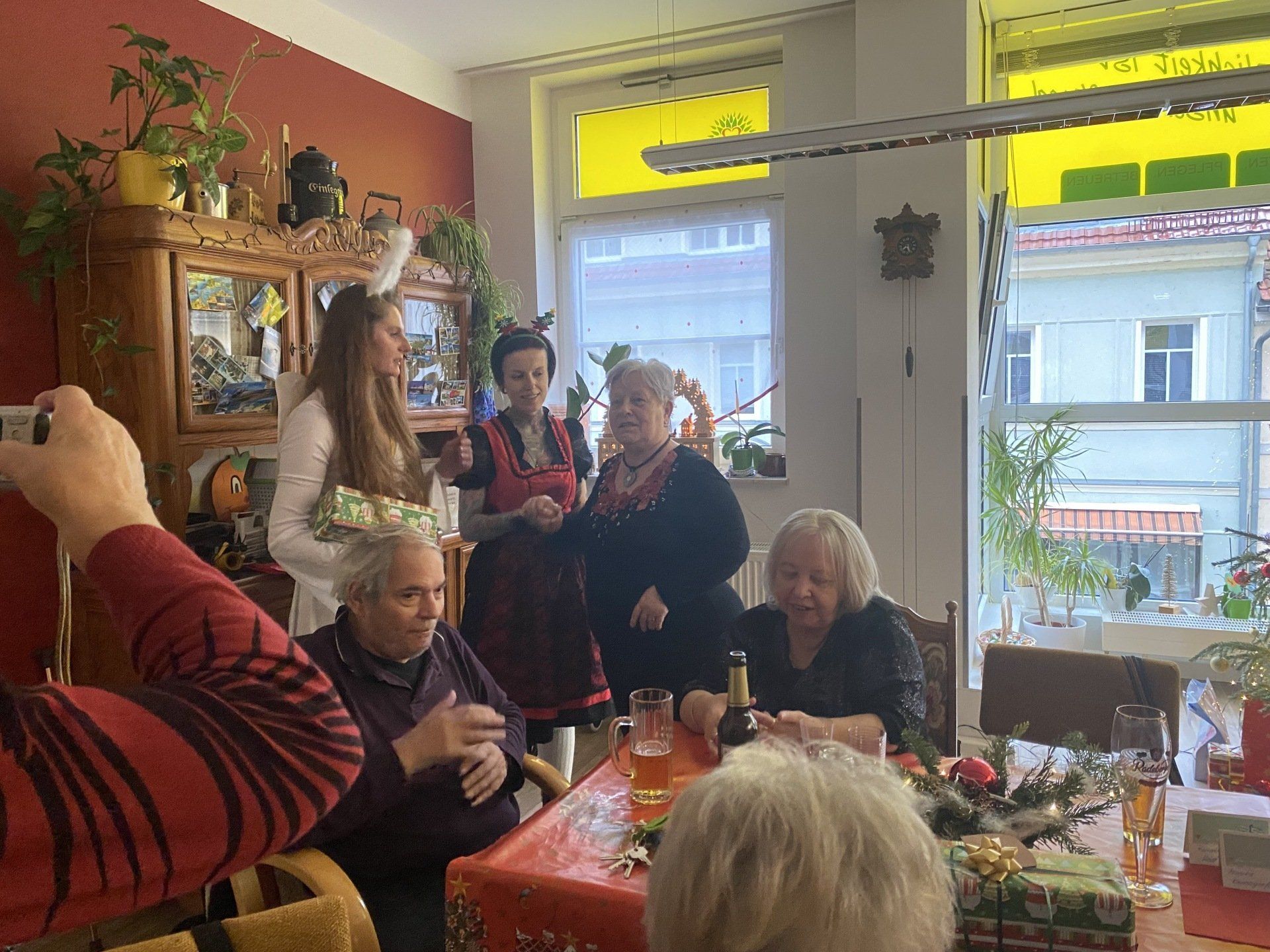 Menschen versammelten sich um einen Tisch mit Geschenken. Im Hintergrund ein Fenster. Weihnachtsdekoration. Einige lächeln.