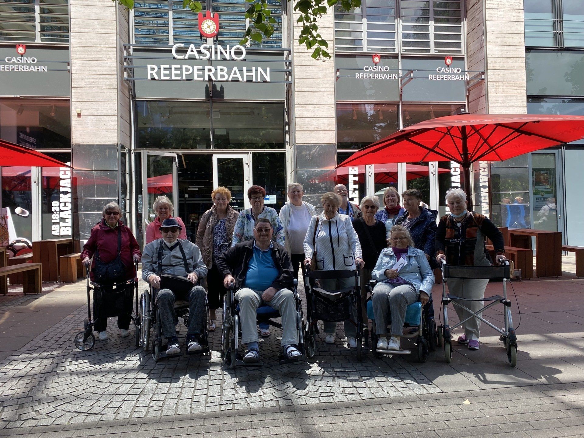 Gruppe von Menschen, einige in Rollstühlen, vor einem Gebäude des „Casino Reeperbahn“. Roter Regenschirm.