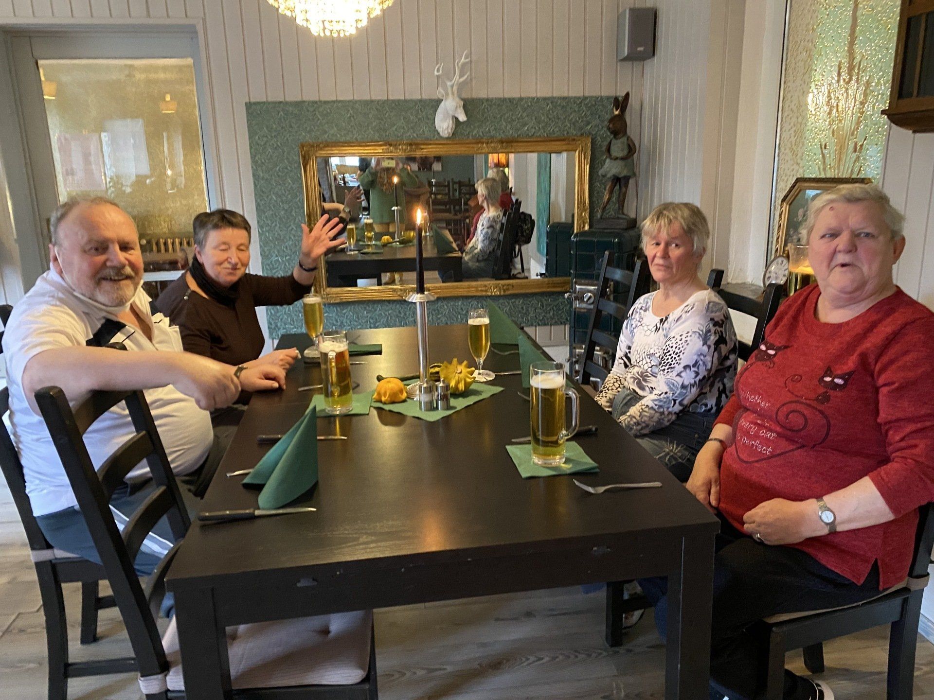 Vier Personen sitzen mit Bier und Essen an einem dunklen Holztisch in einem Restaurant. Einer winkt, andere lächeln.