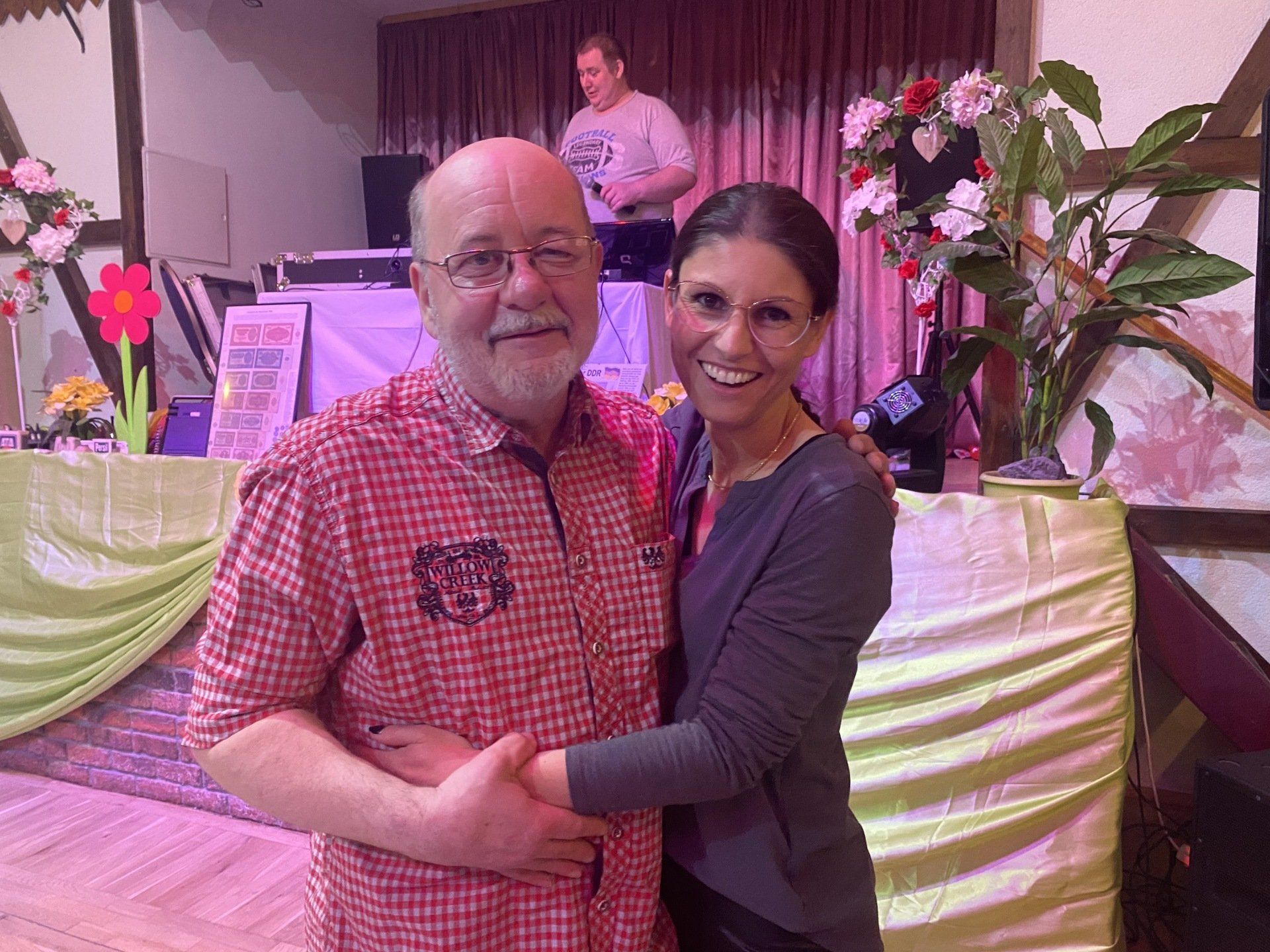 Ein Mann und eine Frau umarmen sich und lächeln in die Kamera, in einem dekorierten Raum mit einem DJ im Hintergrund.